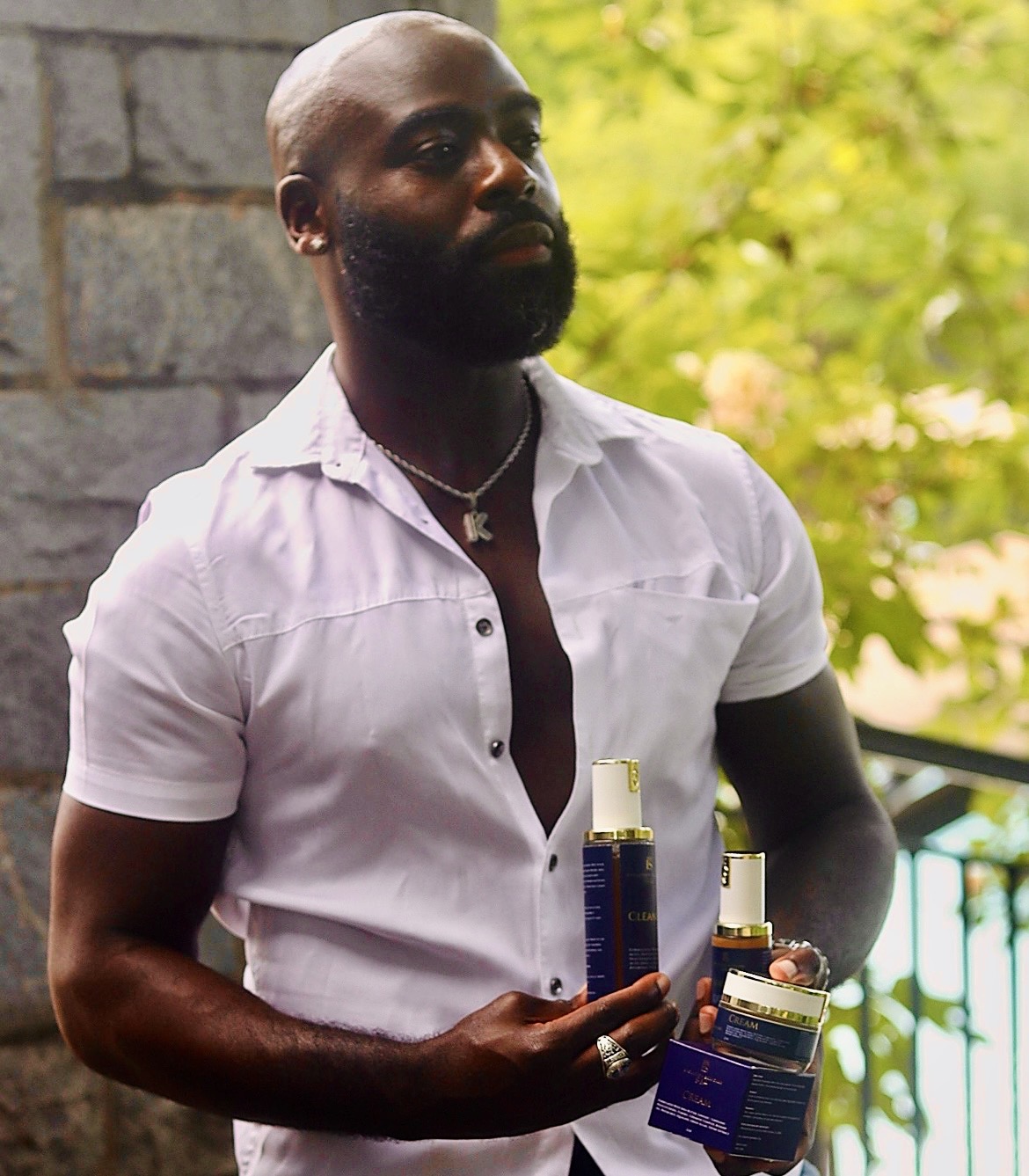 Man with a beard and bald head holding skincare products outdoors, wearing a white shirt, with green foliage in background.