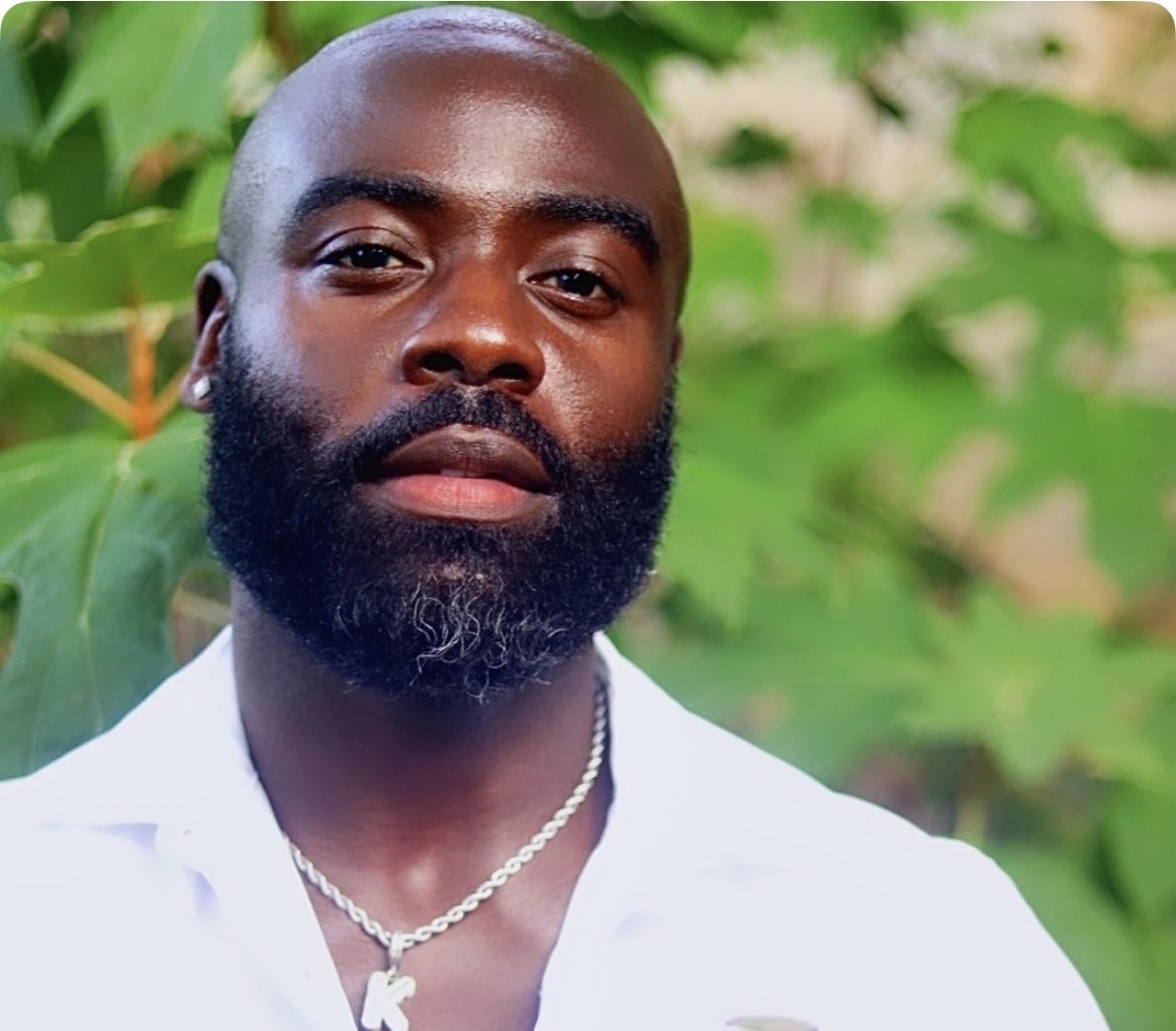 Portrait of a man with a beard and shaved head, wearing a white shirt and a chain necklace, outdoors with green foliage