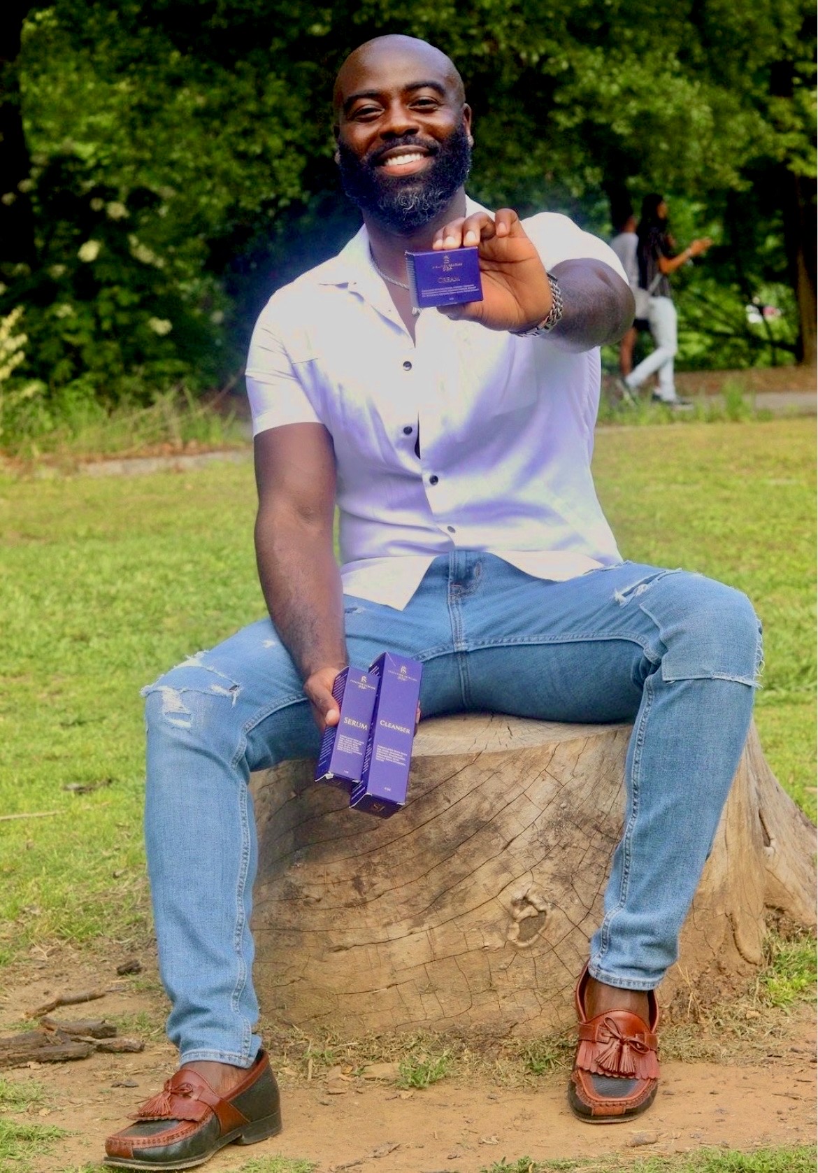 Smiling man sitting on a tree stump outdoors, holding a purple card and a purple box, with green trees in background.