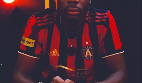 Young man sitting with hands clasped, wearing a red and black sports jersey, in a dimly lit room with a red light in the background.