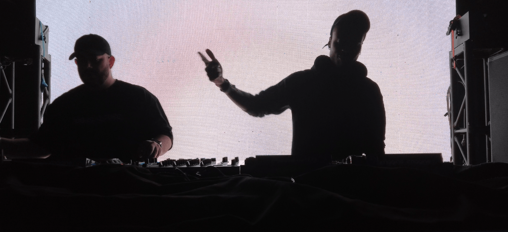 Two DJs perform on stage with a bright background, one making a peace sign gesture, both in silhouette.