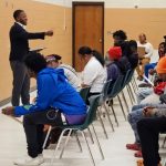 Person standing and speaking to seated group in a classroom or seminar setting.