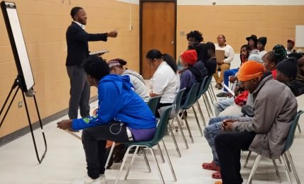 Person standing and speaking to seated group in a classroom or seminar setting.