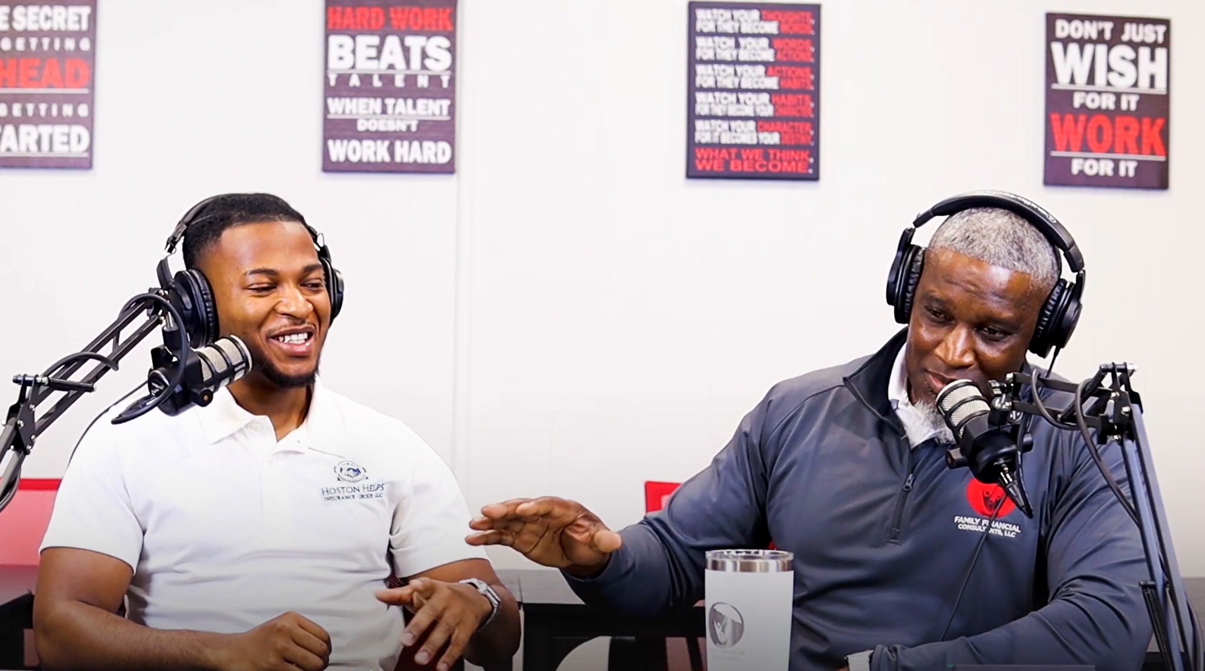 Two people wearing headphones sit at microphones in a recording studio, smiling and engaging in conversation.