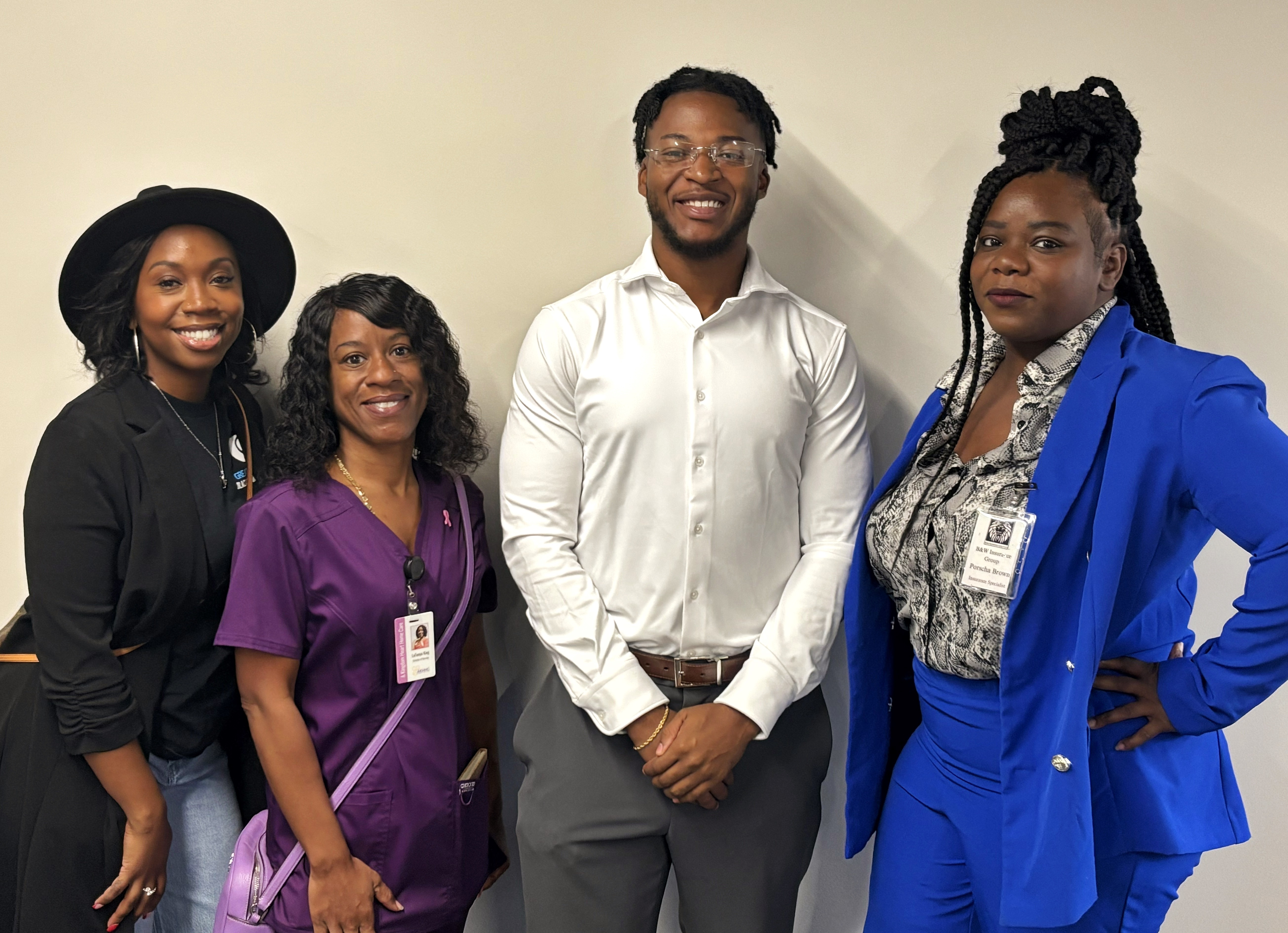 Four people standing together, smiling, against a plain background. One woman wears a black hat and jacket, another in purple scrubs, a man in a white shirt, and a woman in a blue suit.