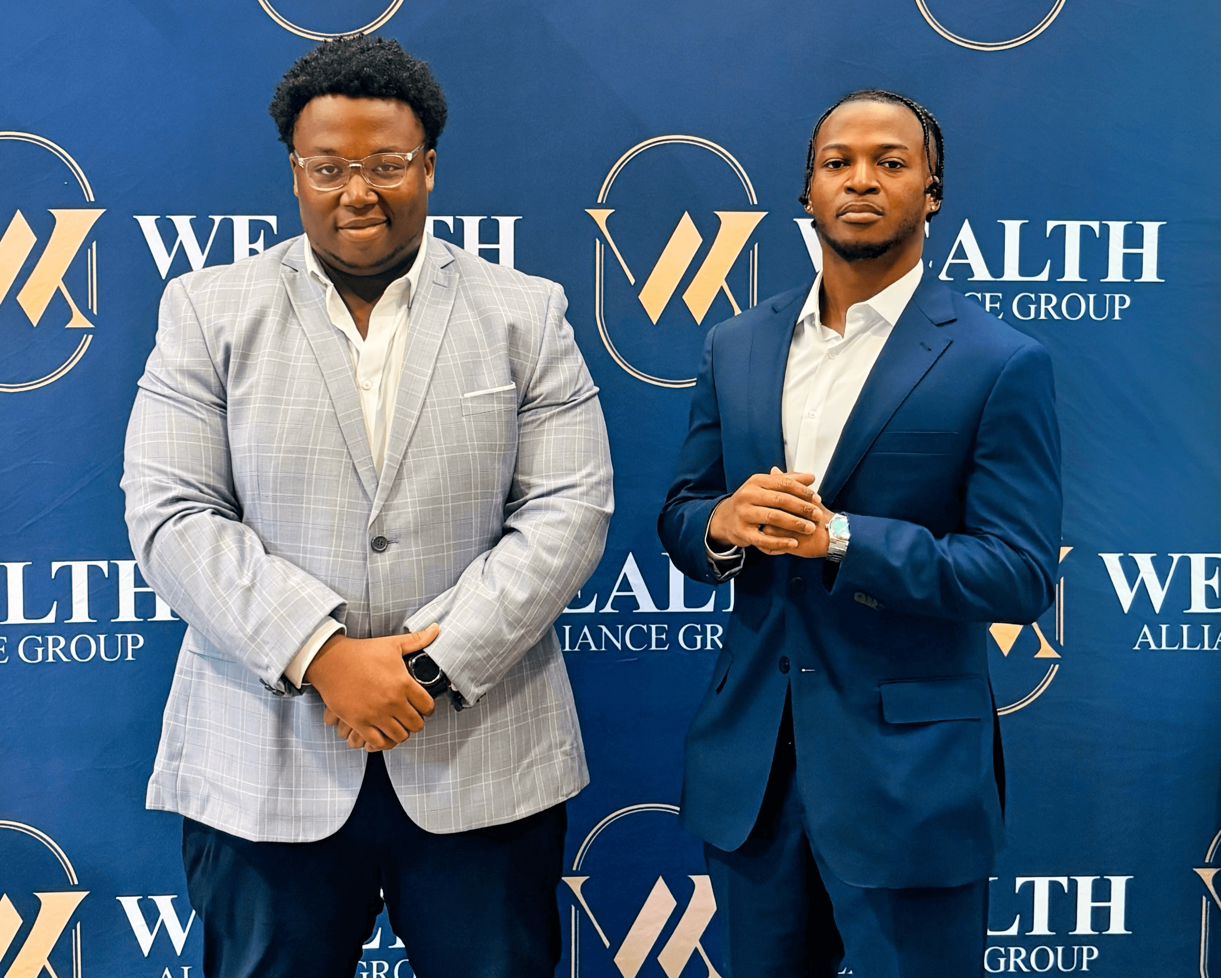 Two men in suits standing in front of a blue backdrop with white and gold text and logo, smiling at the camera.
