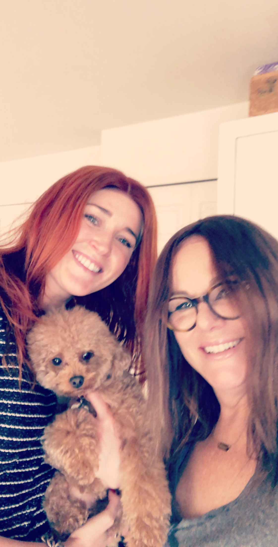 Two women smiling, one holding a small curly-haired dog, in an indoor setting.
