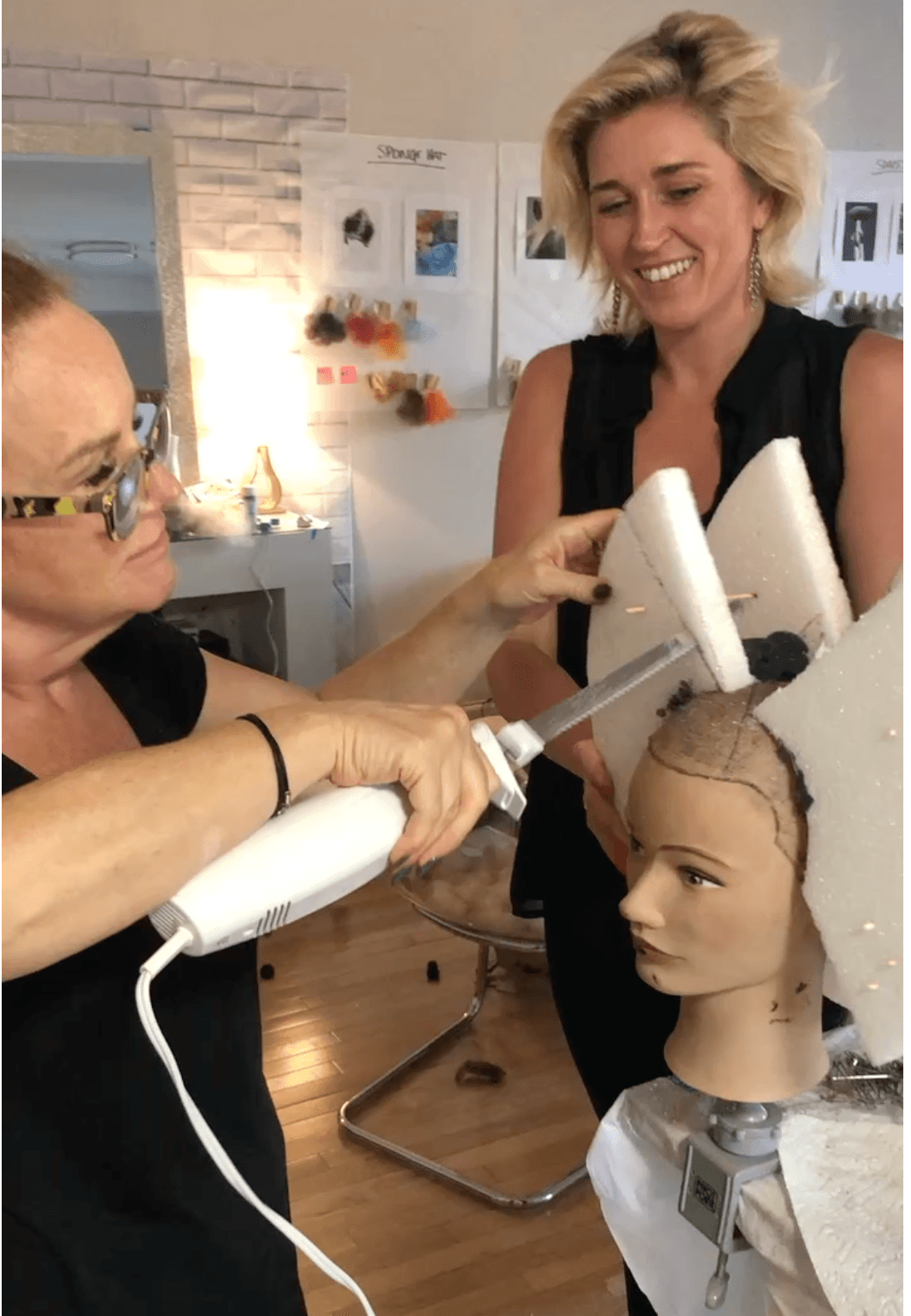 Woman with short blonde hair smiling at a mannequin head with hairdressing tools, in a salon setting.