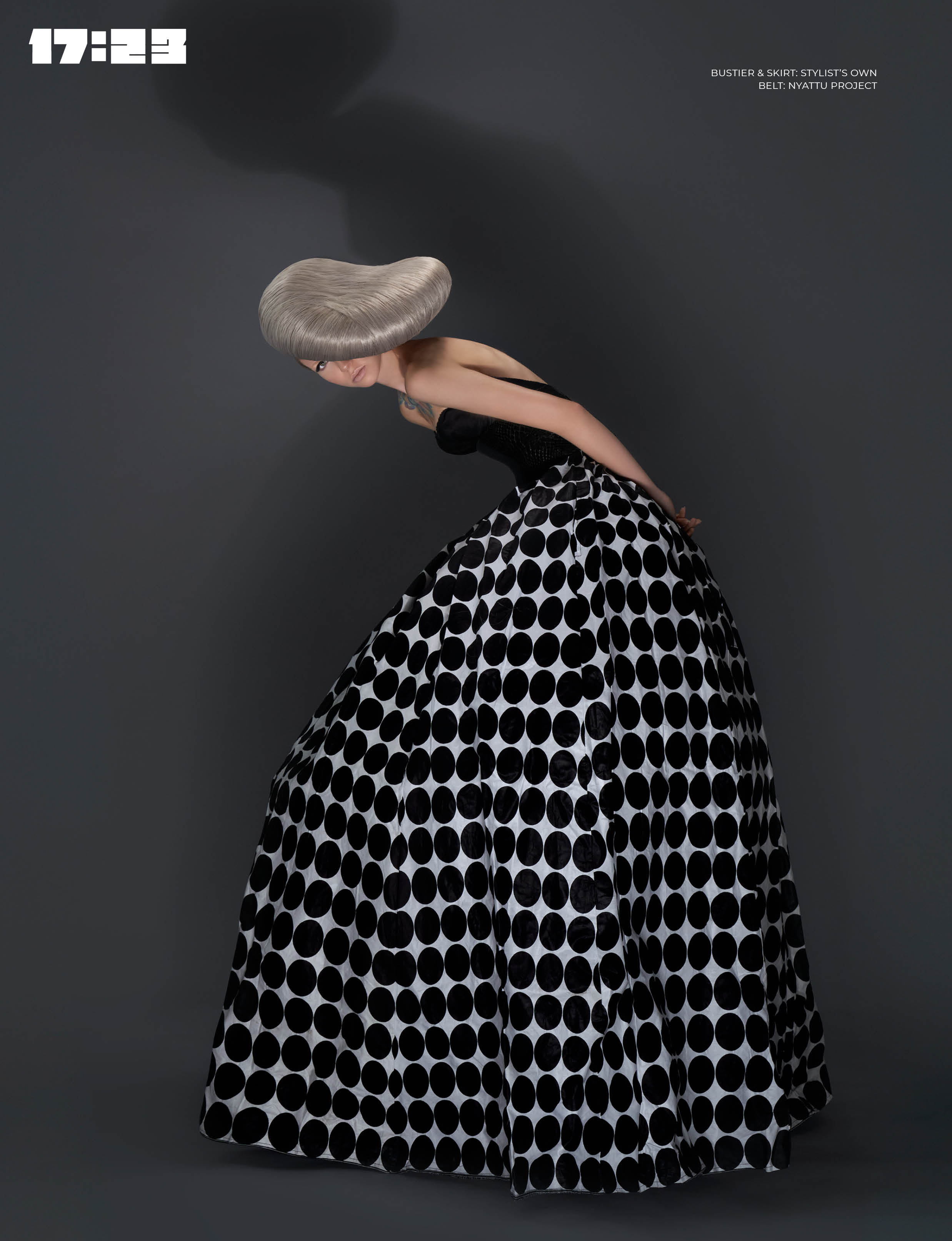 Person with a large, wide-brimmed hat bending forward, wearing a polka dot skirt, against a dark background.