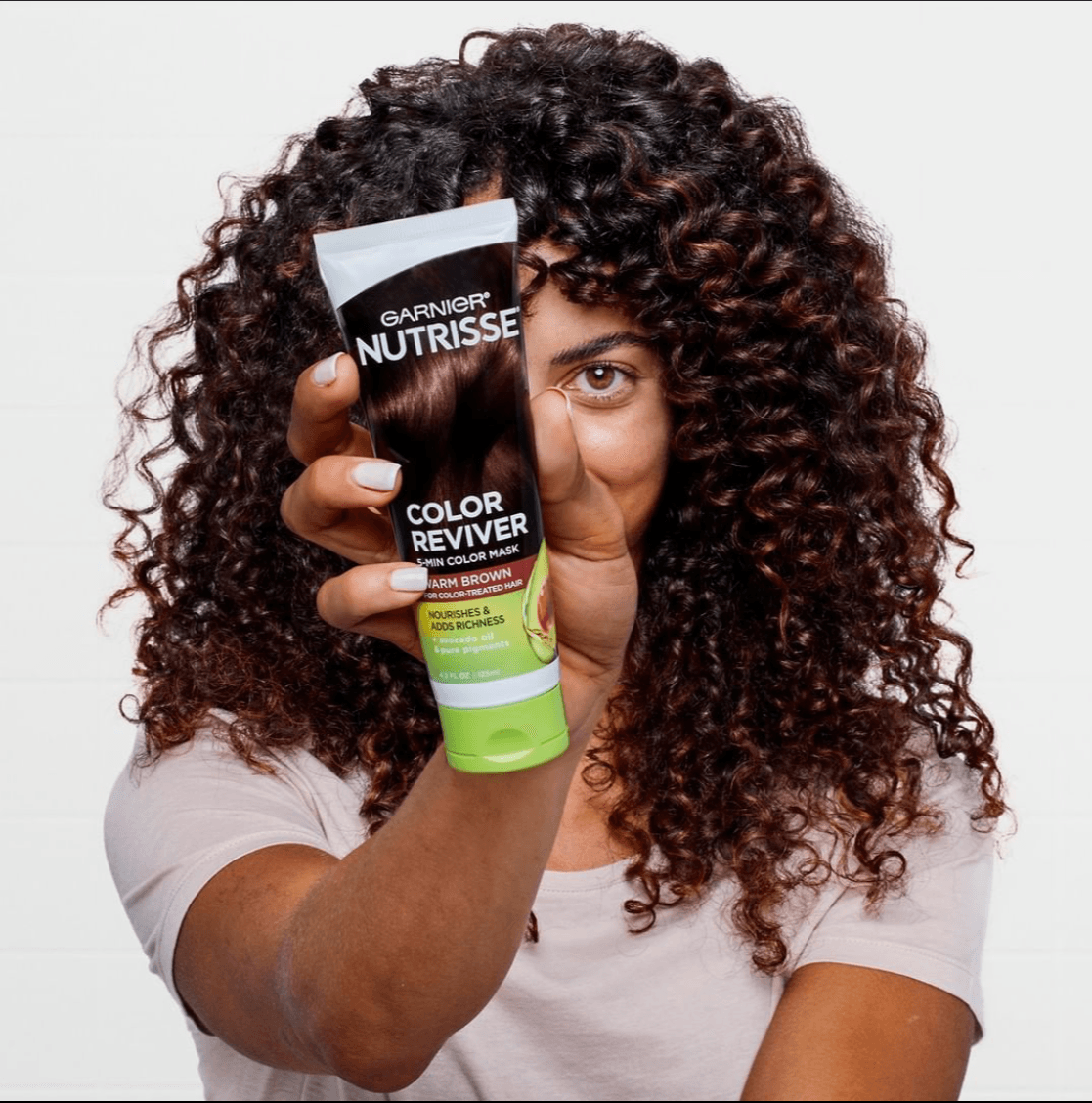 Woman with curly hair holding Garnier NutriSse Color Reviver tube, partially covering her face, smiling, white background.