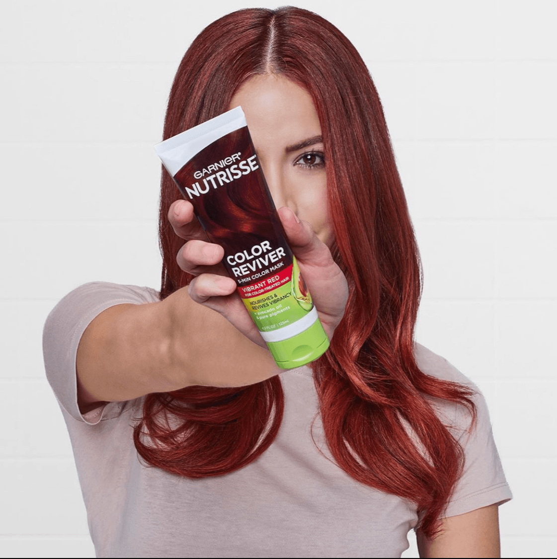 Woman with long red hair holding a hair color product tube in front of her face, partially covering it, against a white background.