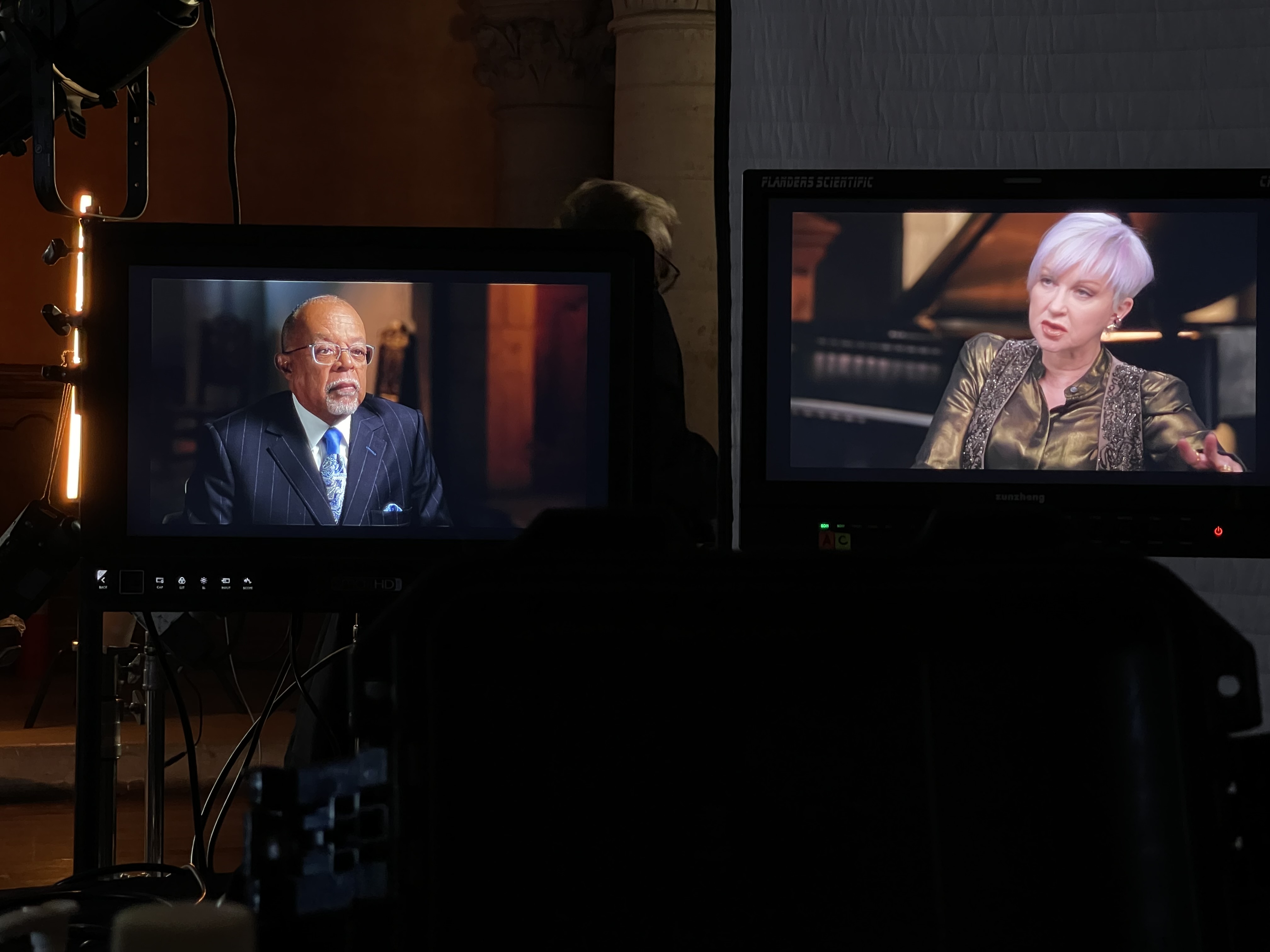 Two monitors display people in interview settings, one with a man and the other with a woman, in a studio environment.