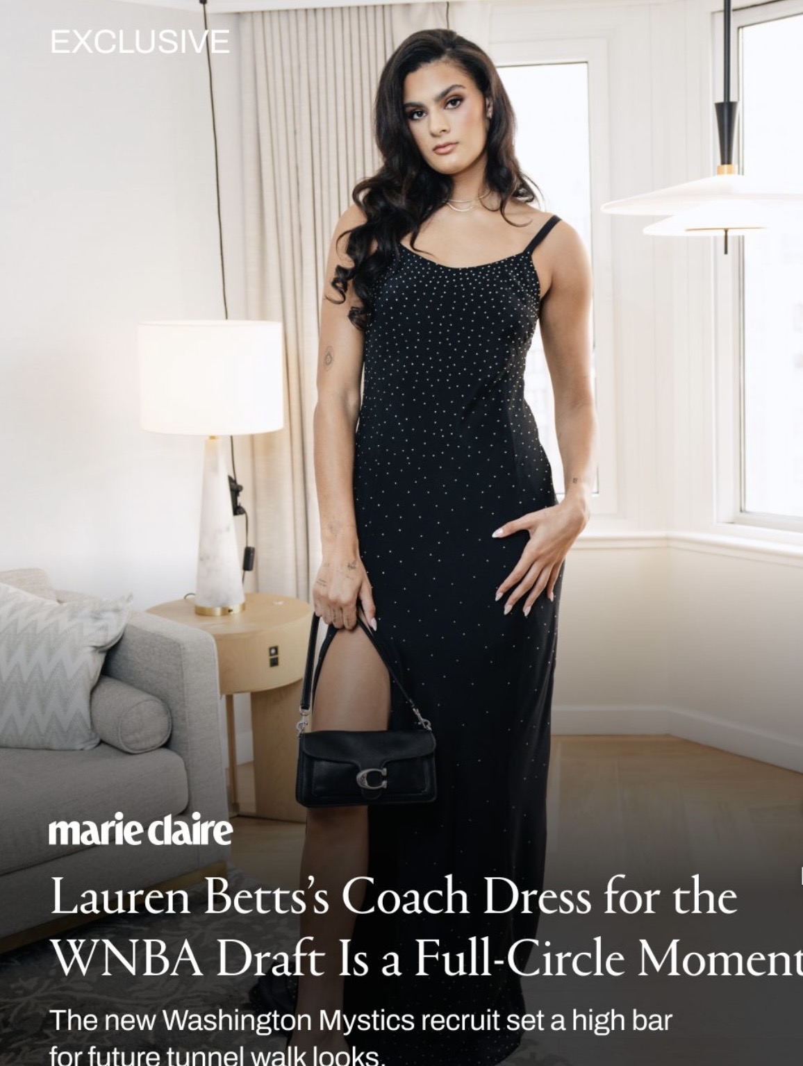 Woman in a black dress with polka dots holding a black handbag in a well-lit room with a sofa and lamp.