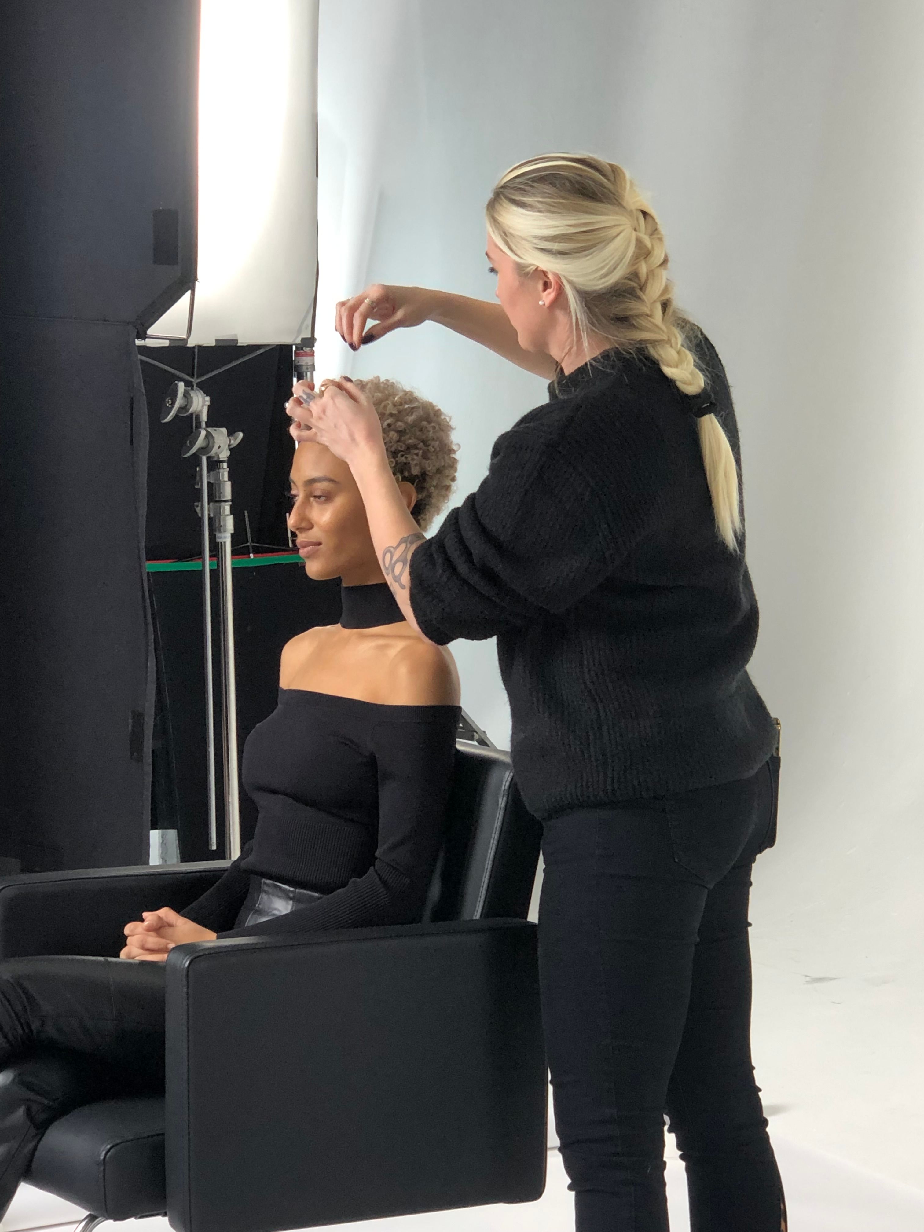 A woman with blonde hair styled in a braid is applying makeup to a seated woman with curly hair, in a studio setting.