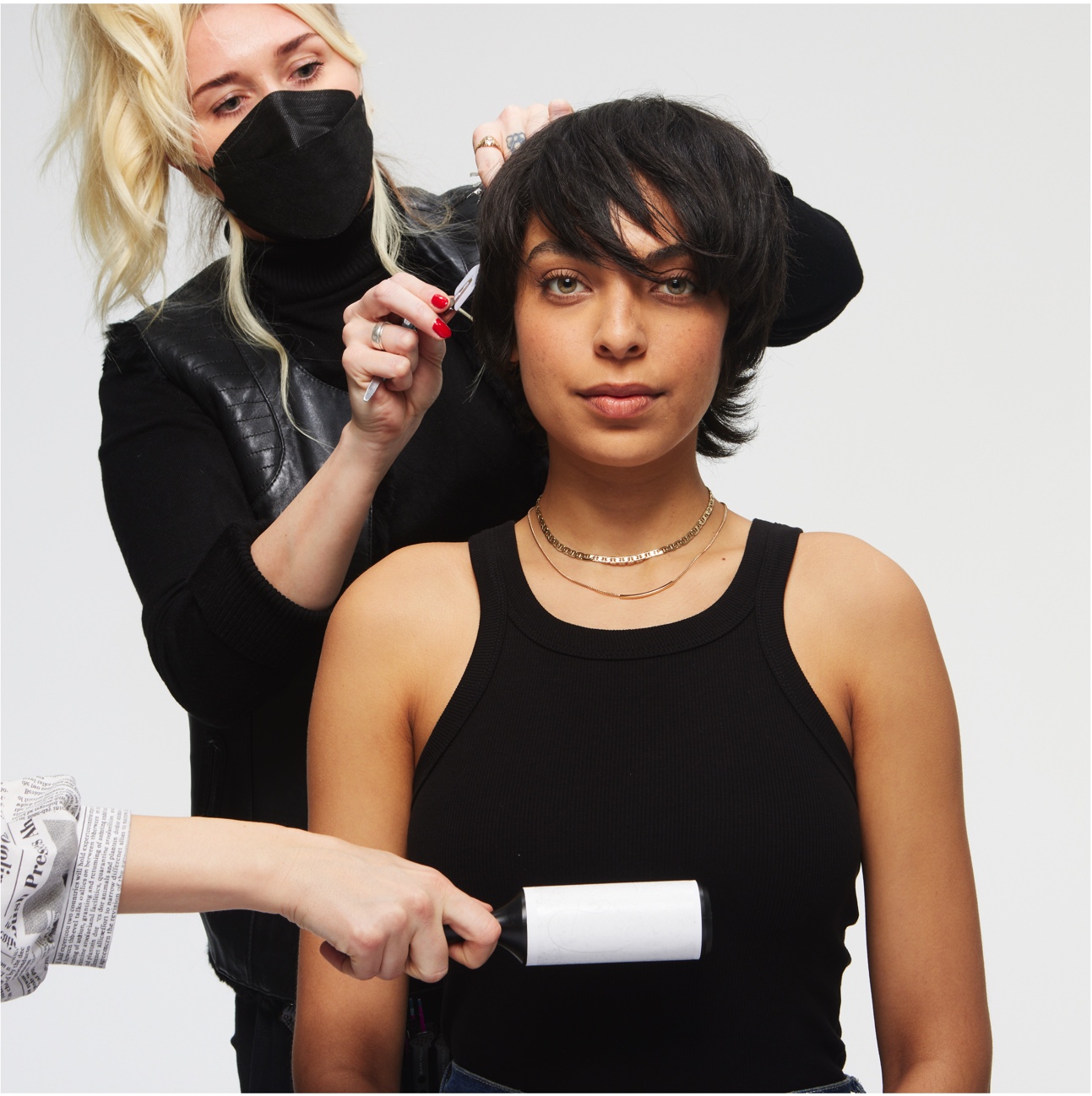 Woman with short dark hair in a black tank top getting her hair styled by a stylist with blonde hair, black mask, and black outfit, using a curling iron.