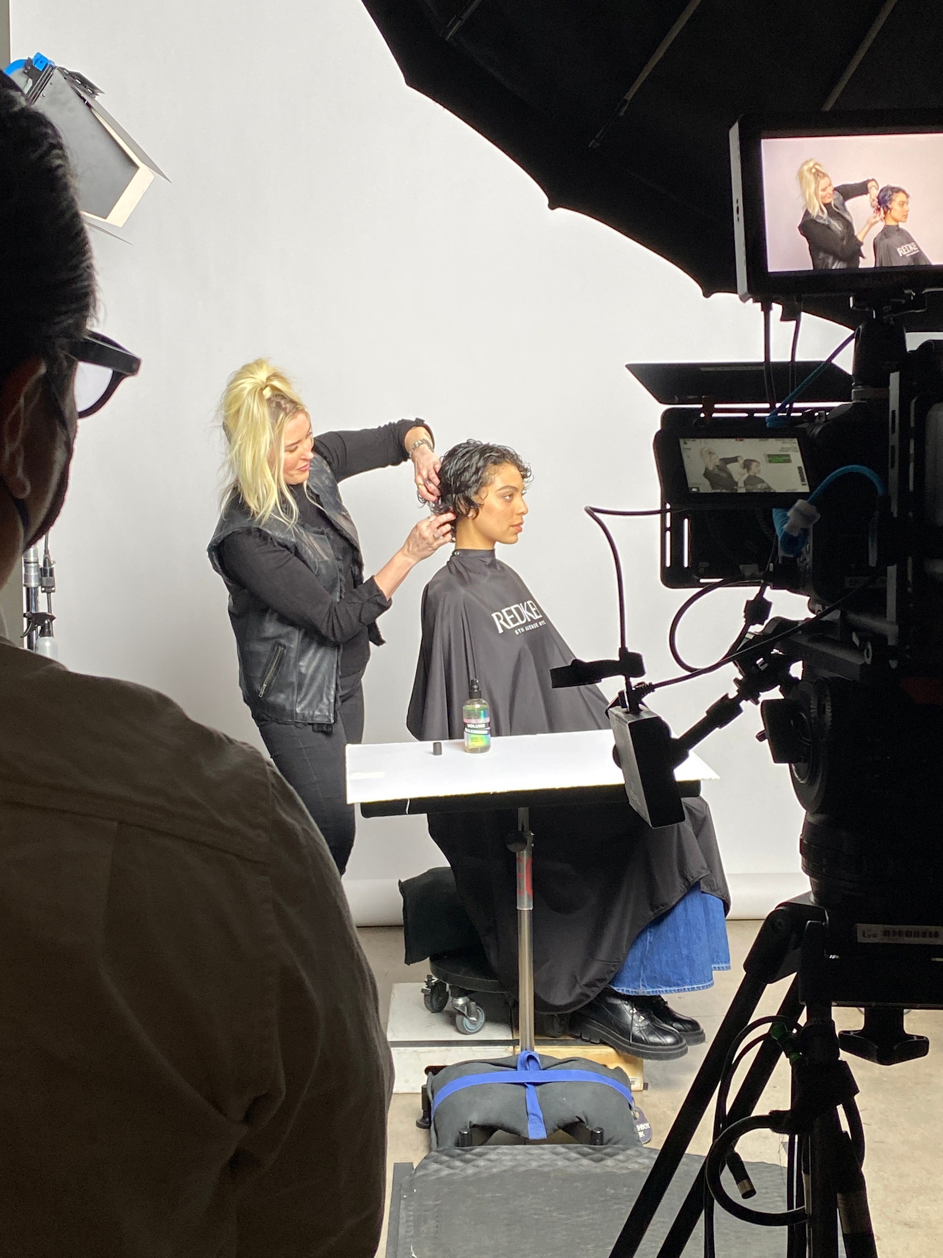 Hairdresser styles a person's hair in a studio with filming equipment and a white background.