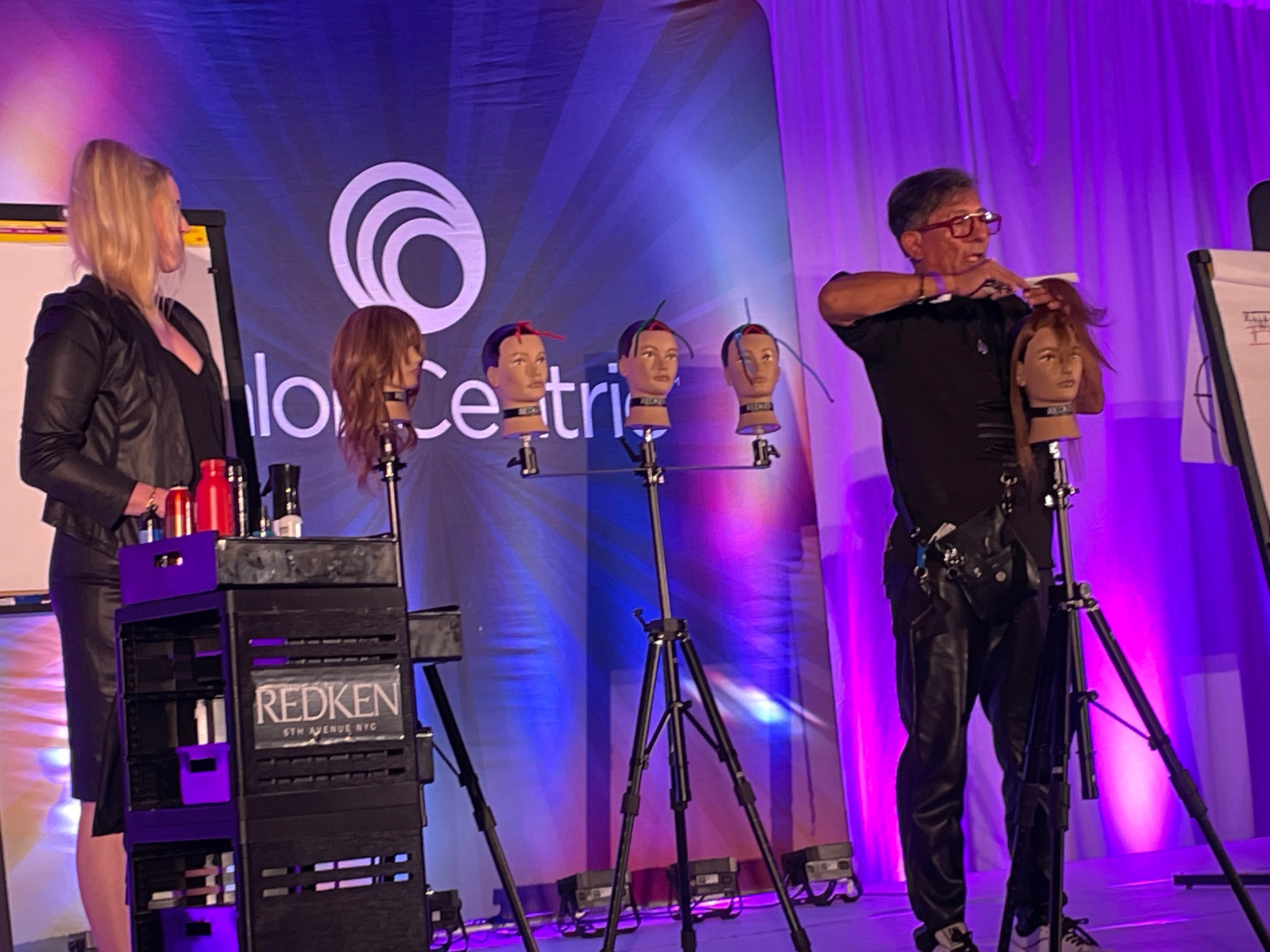 Hairdressing demonstration with a stylist working on a mannequin head, audience watching, colorful stage background, hair products on a cart.