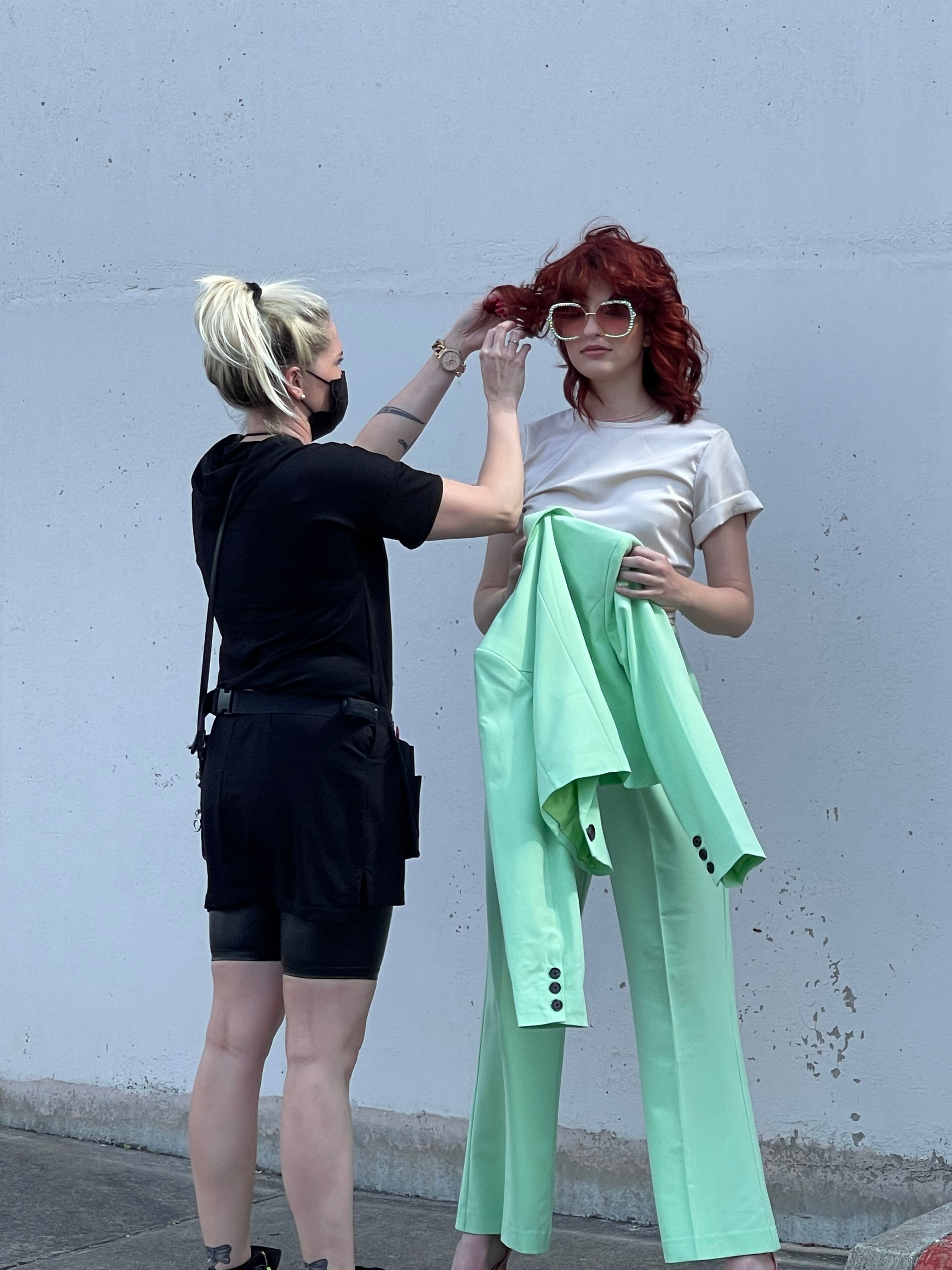 Two women stand against a plain wall, one adjusting the other's hair, the other holding a green jacket and wearing sunglasses.