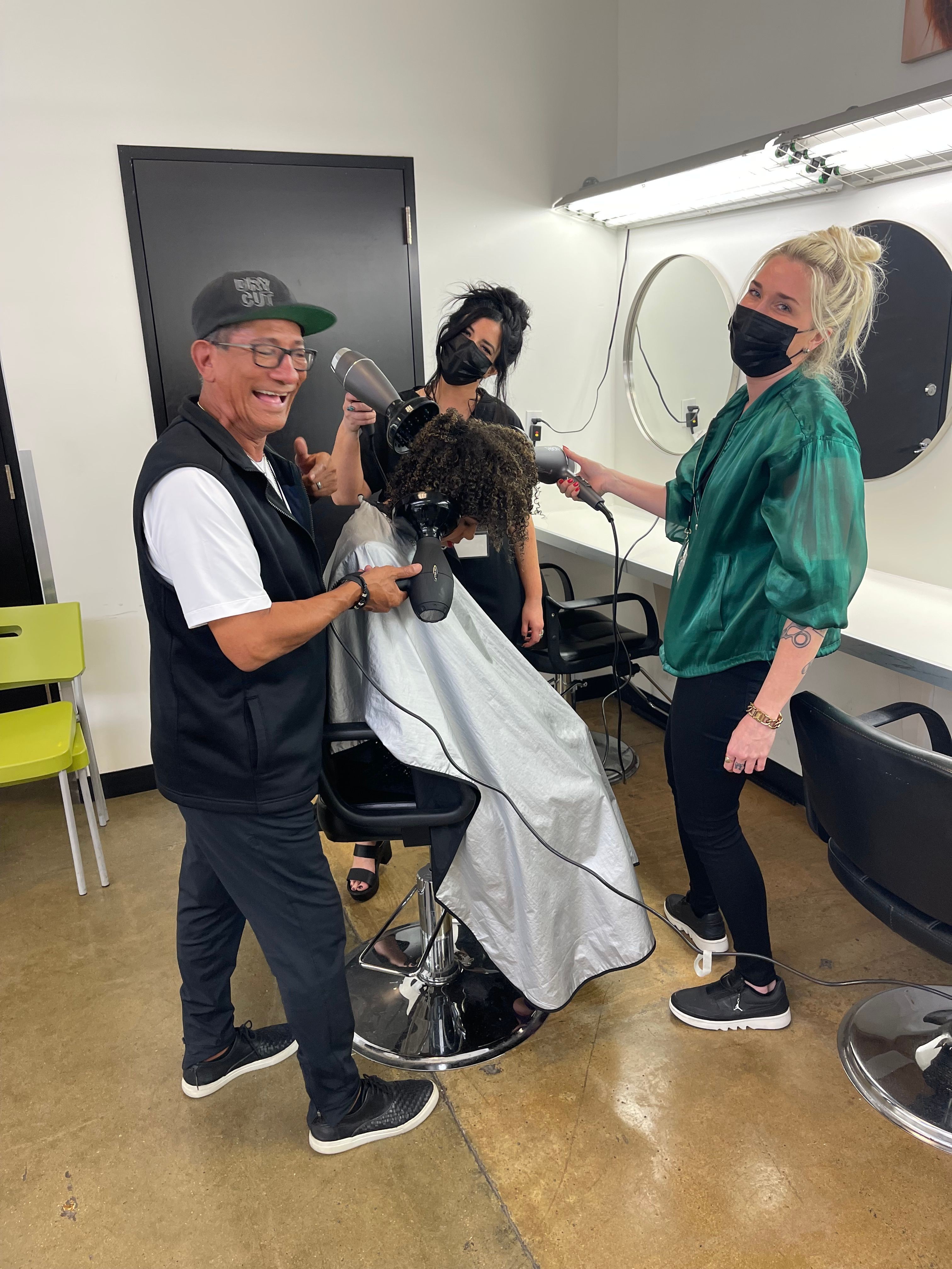 Person getting a haircut in a salon, with two staff members attending, one holding scissors and the other with a cape, all wearing masks.