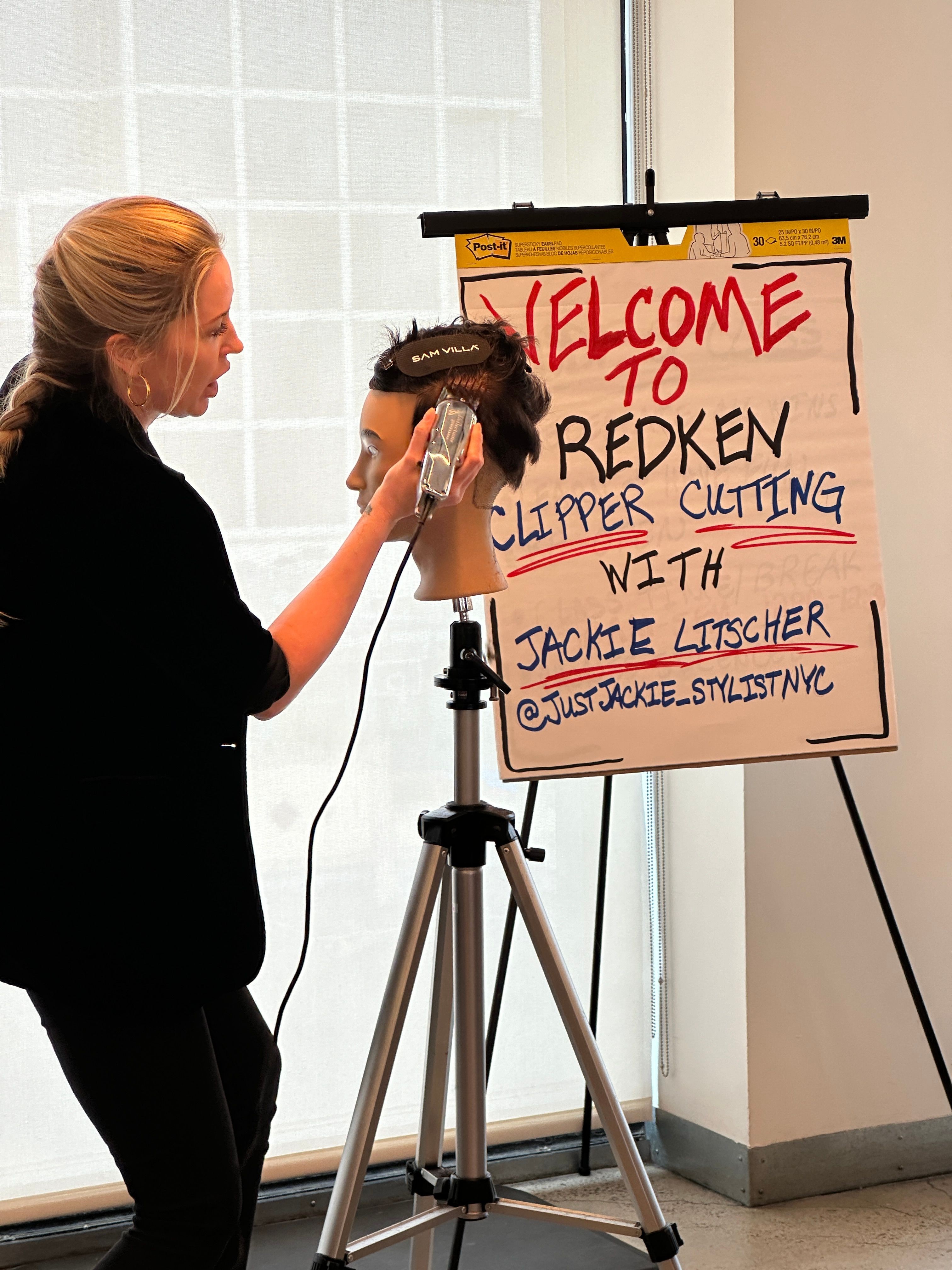 Woman styling a person's hair in front of a welcome sign for Redken event, with tripod and window in background.