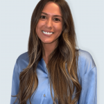 Smiling woman with long wavy hair wearing a blue shirt, facing forward, against a light background.