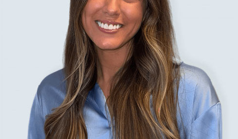 Smiling woman with long wavy hair wearing a blue shirt, facing forward, against a light background.