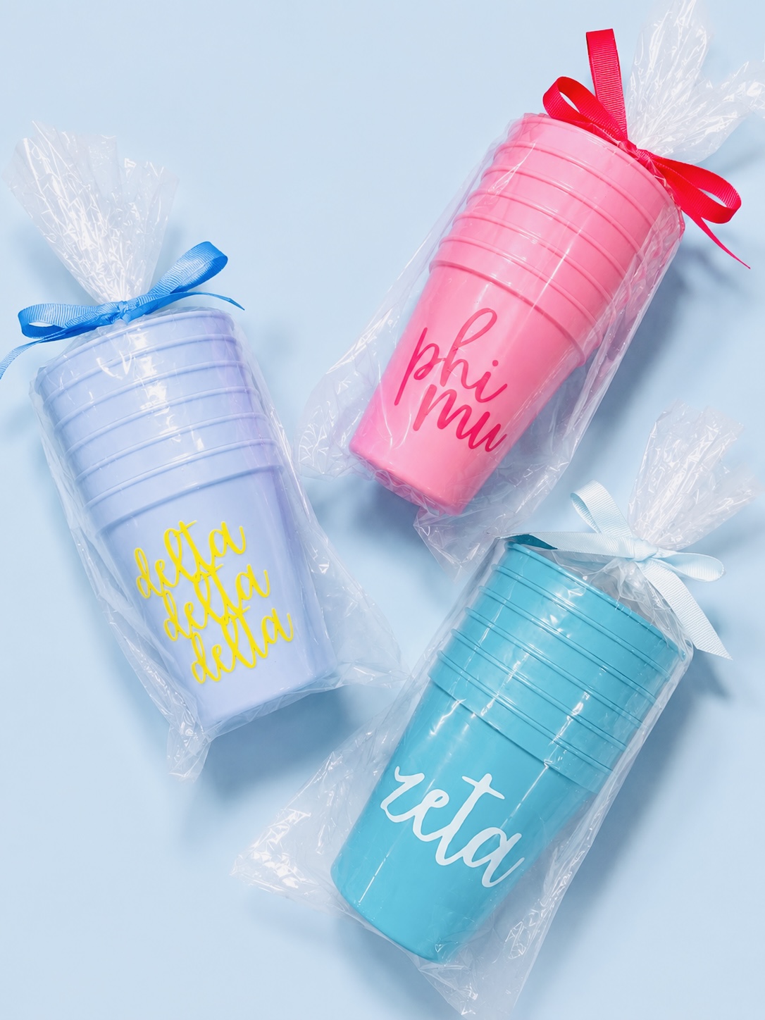 Three colorful cylindrical containers with ribbons, one pink, one blue, and one purple, each with text and wrapped in plastic.