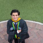 Young man in graduation gown and medals smiling with arms crossed, standing on brick pathway with grass background.