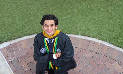 Young man in graduation gown and medals smiling with arms crossed, standing on brick pathway with grass background.