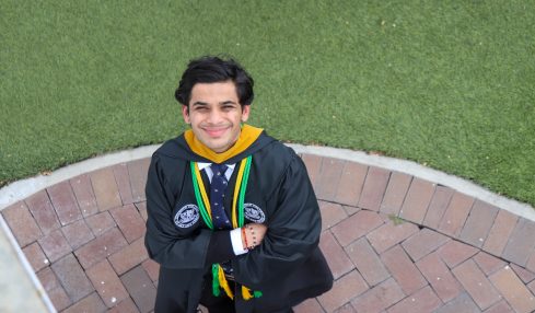 Young man in graduation gown and medals smiling with arms crossed, standing on brick pathway with grass background.