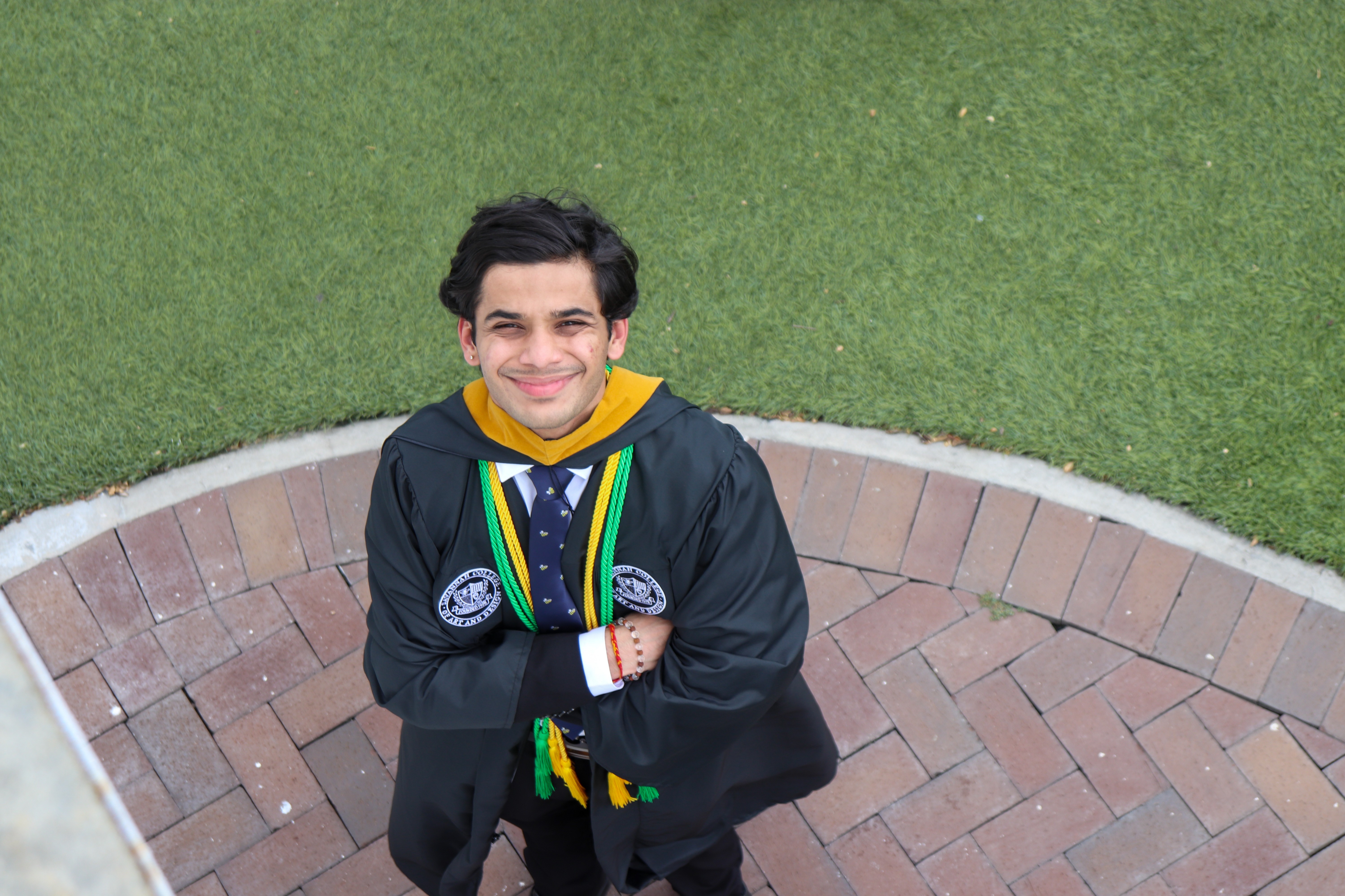 Young man in graduation gown and medals smiling with arms crossed, standing on brick pathway with grass background.