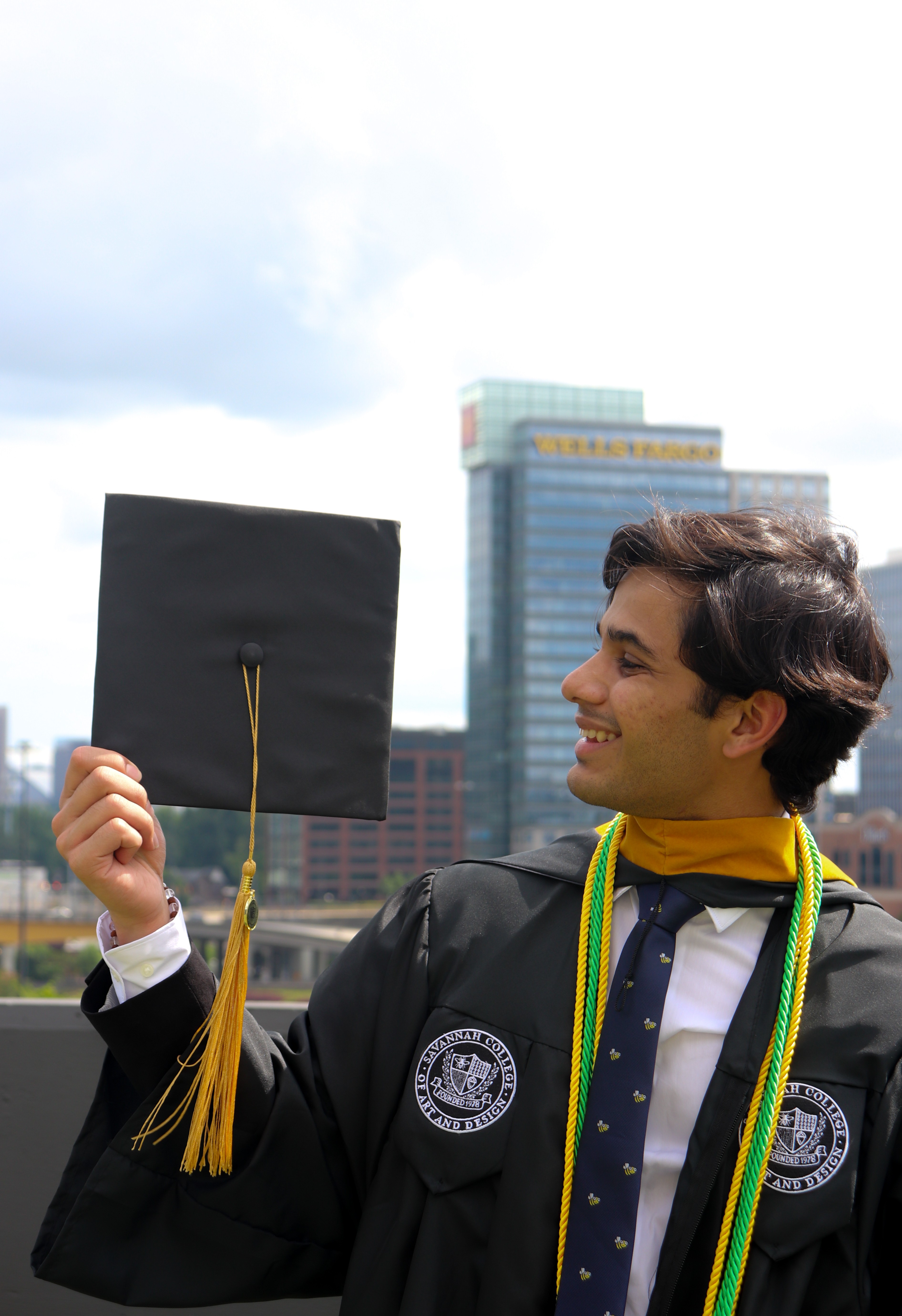 Young man in graduation gown holding a cap outdoors with city buildings in background, smiling, facing right.
