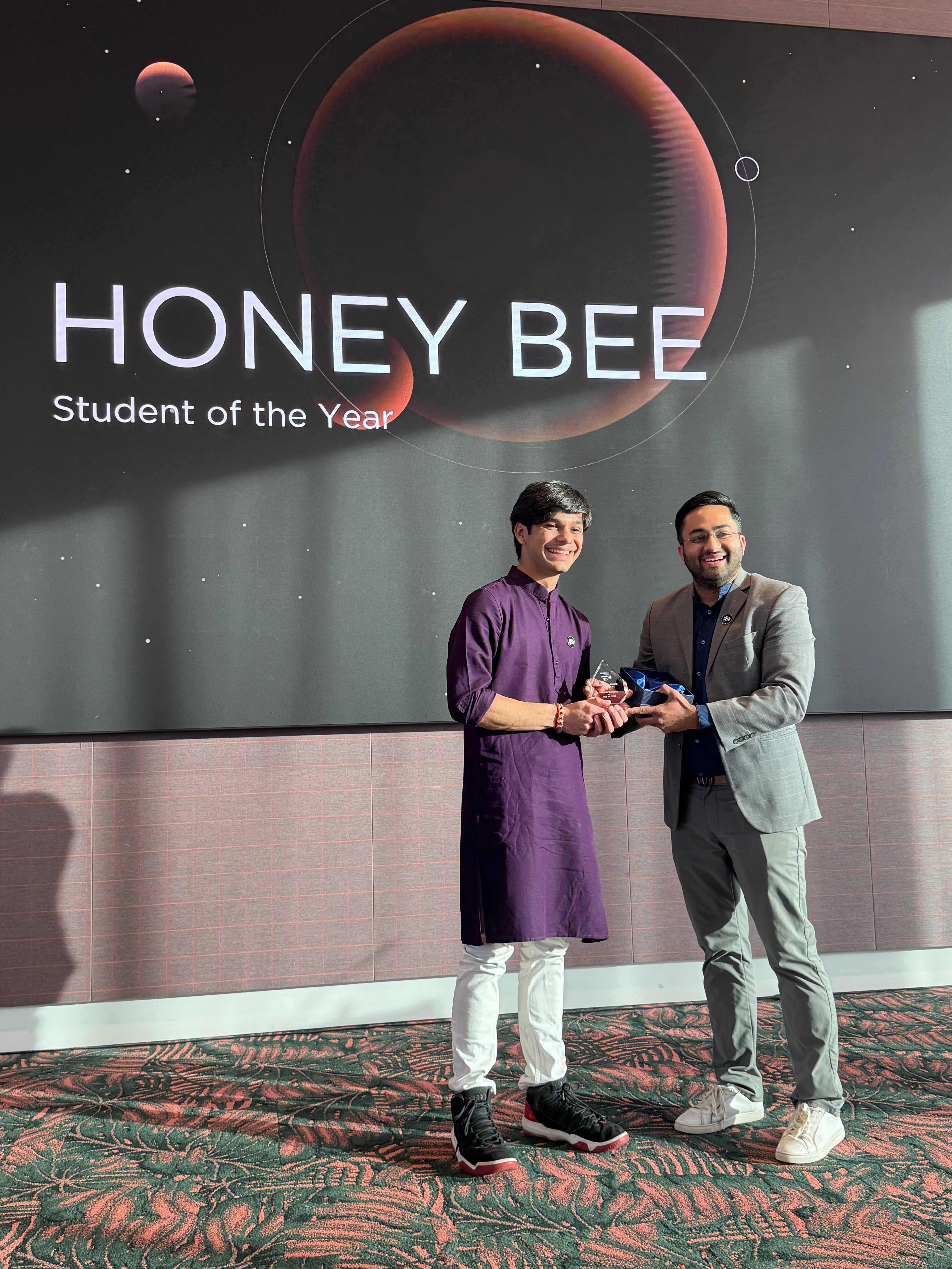 Two people standing on stage, one handing an award to the other, with a large screen behind them displaying the event name.