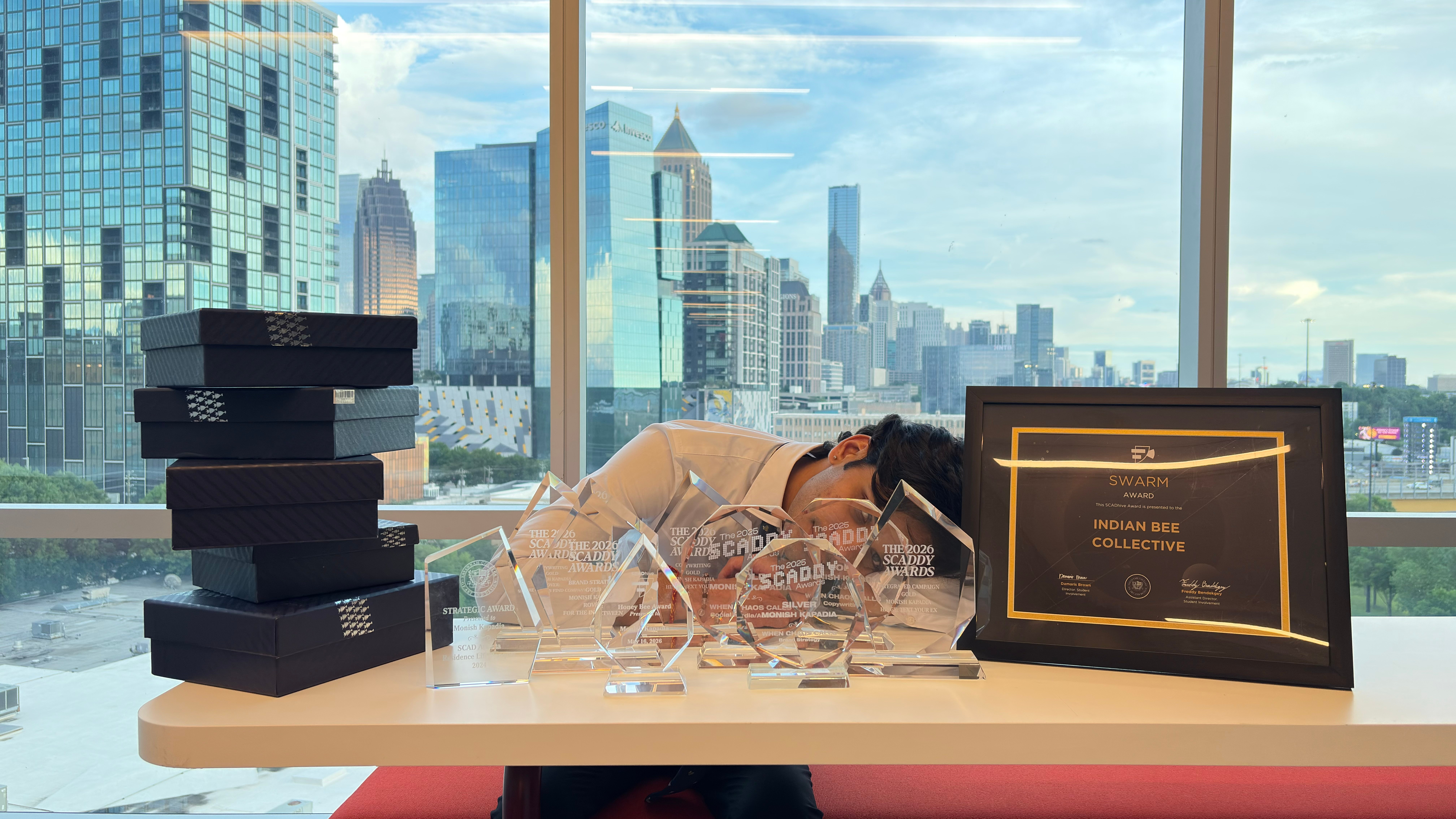 Person resting head on desk with awards and a framed certificate, city skyline visible through large window behind.