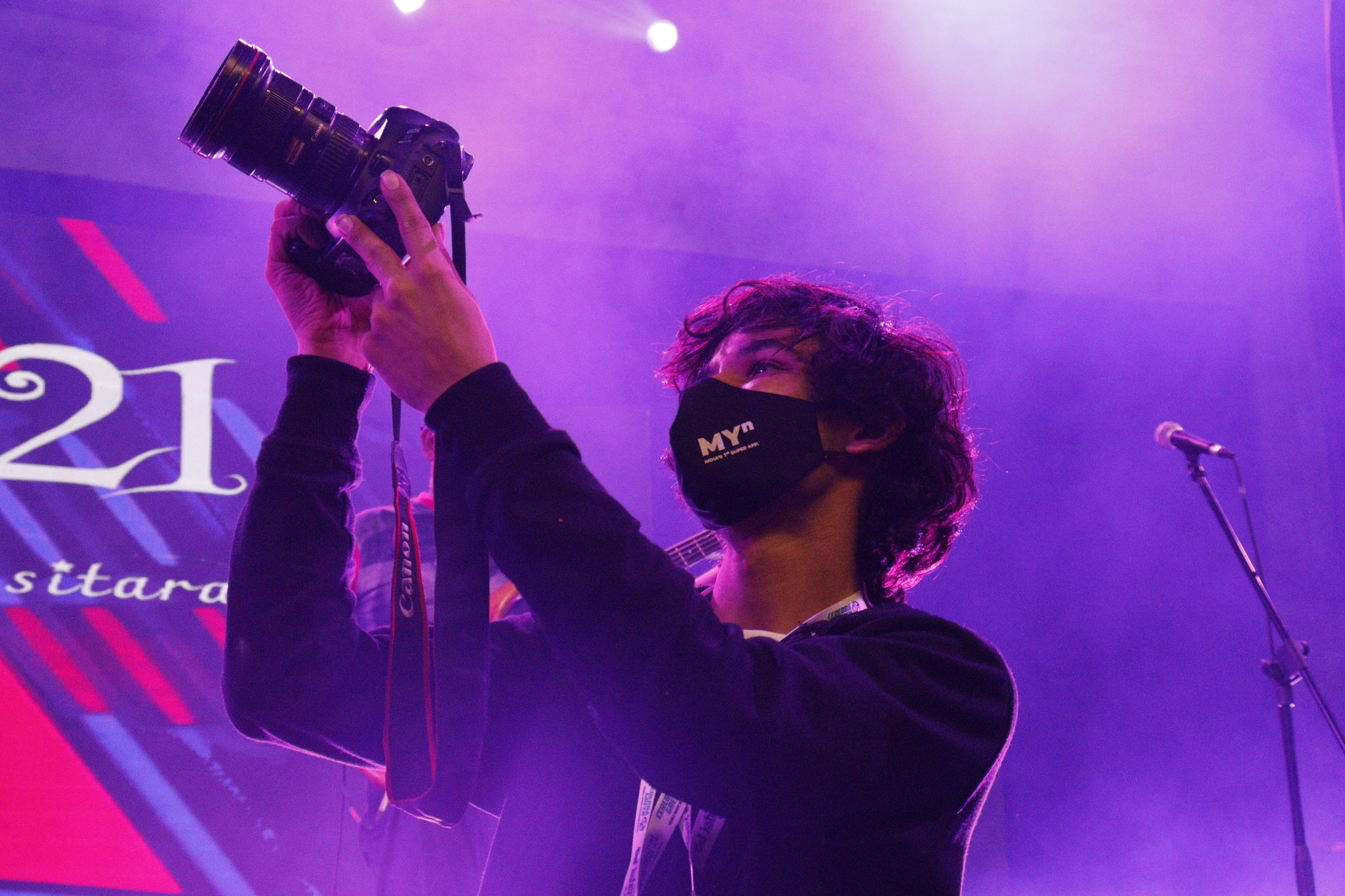 Person with curly hair wearing a face mask holding a camera on stage with purple lighting, microphone stand nearby.
