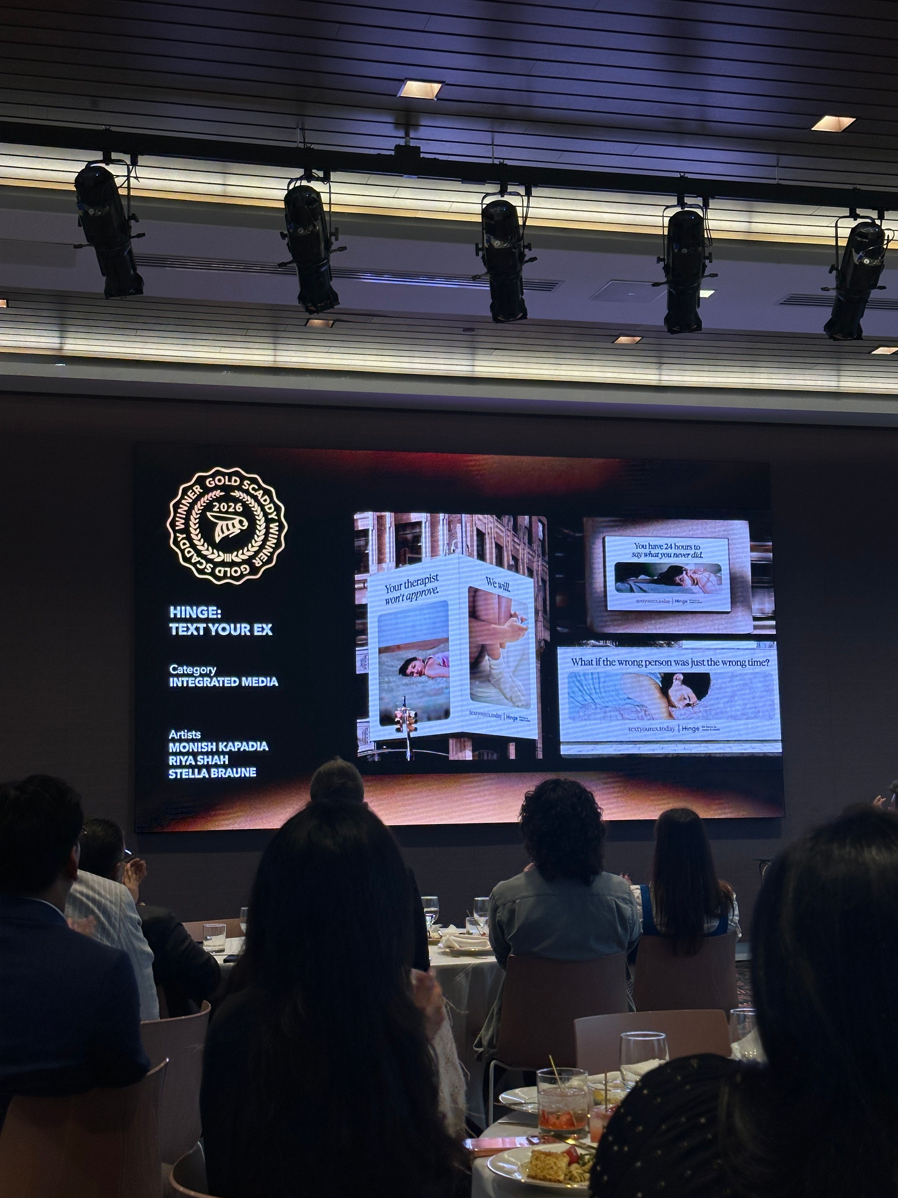 Audience watching a presentation with slides on a large screen, showing images and text, in a dark room.