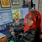 Person with red hair and glasses working at a dual-monitor computer setup with art supplies nearby.