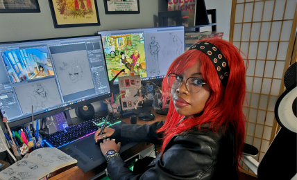Person with red hair and glasses working at a dual-monitor computer setup with art supplies nearby.