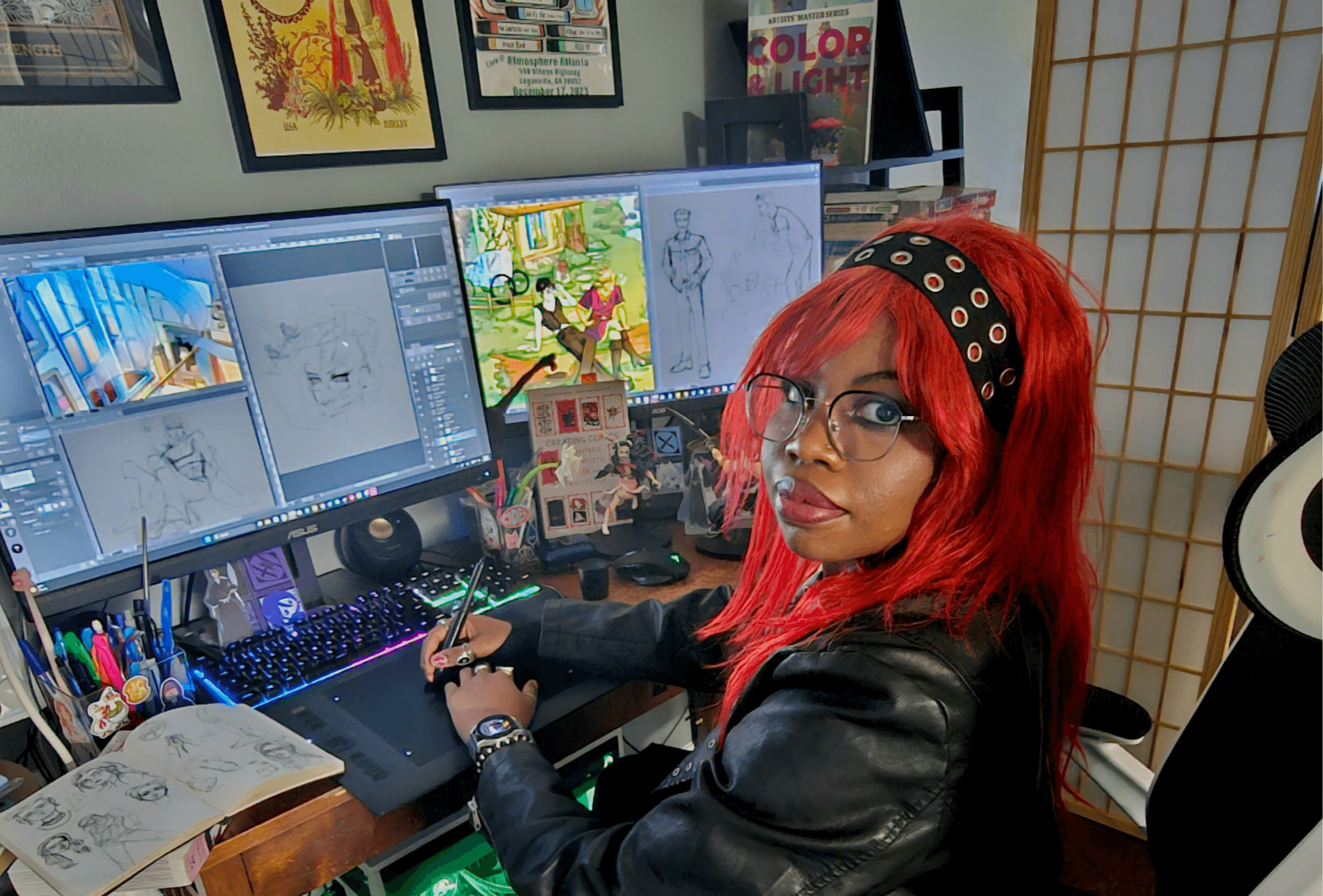 Person with red hair and glasses working at a dual-monitor computer setup with art supplies nearby.