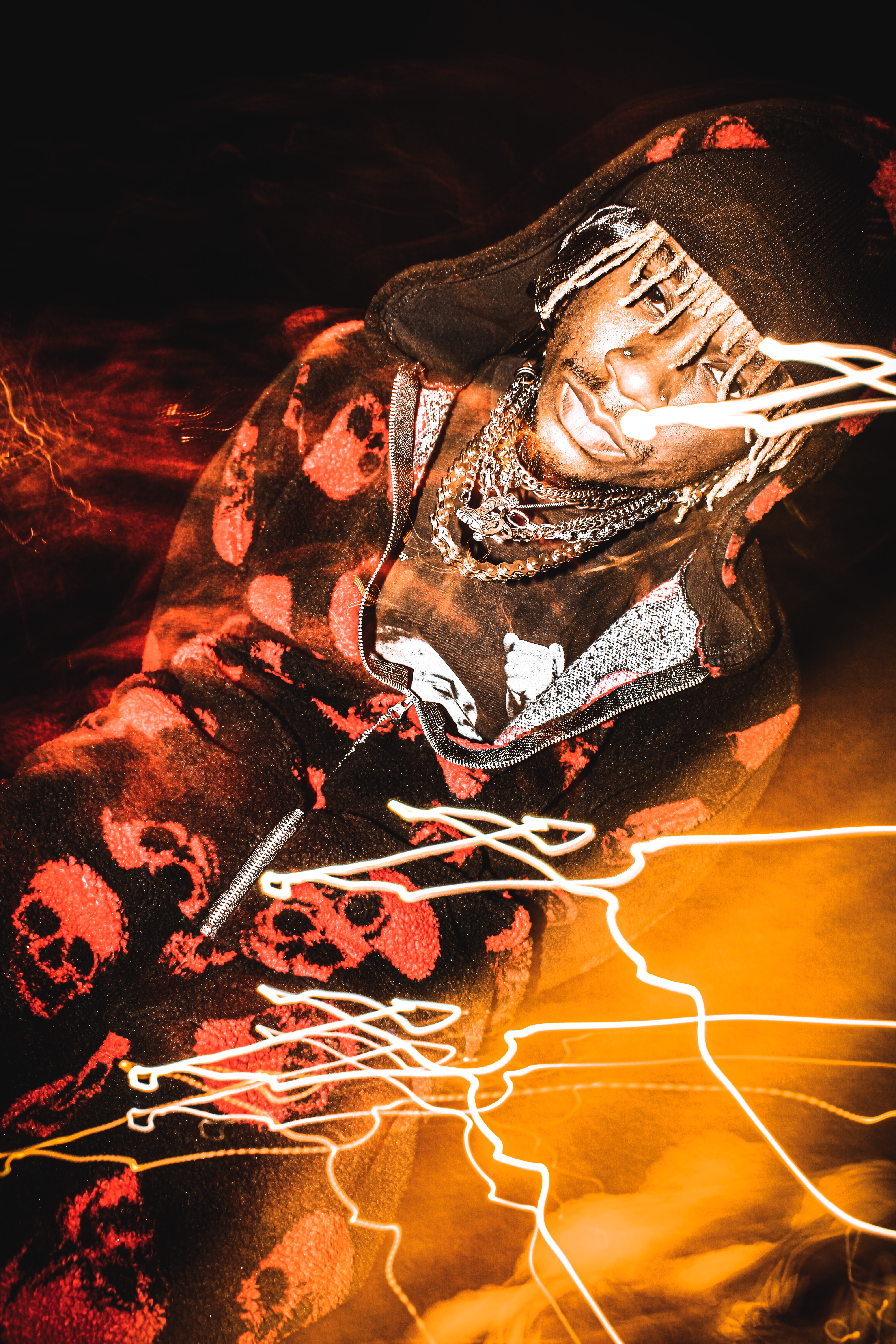 Person wearing a hat and skull-patterned hoodie, with light streaks and fiery background, looking downwards.