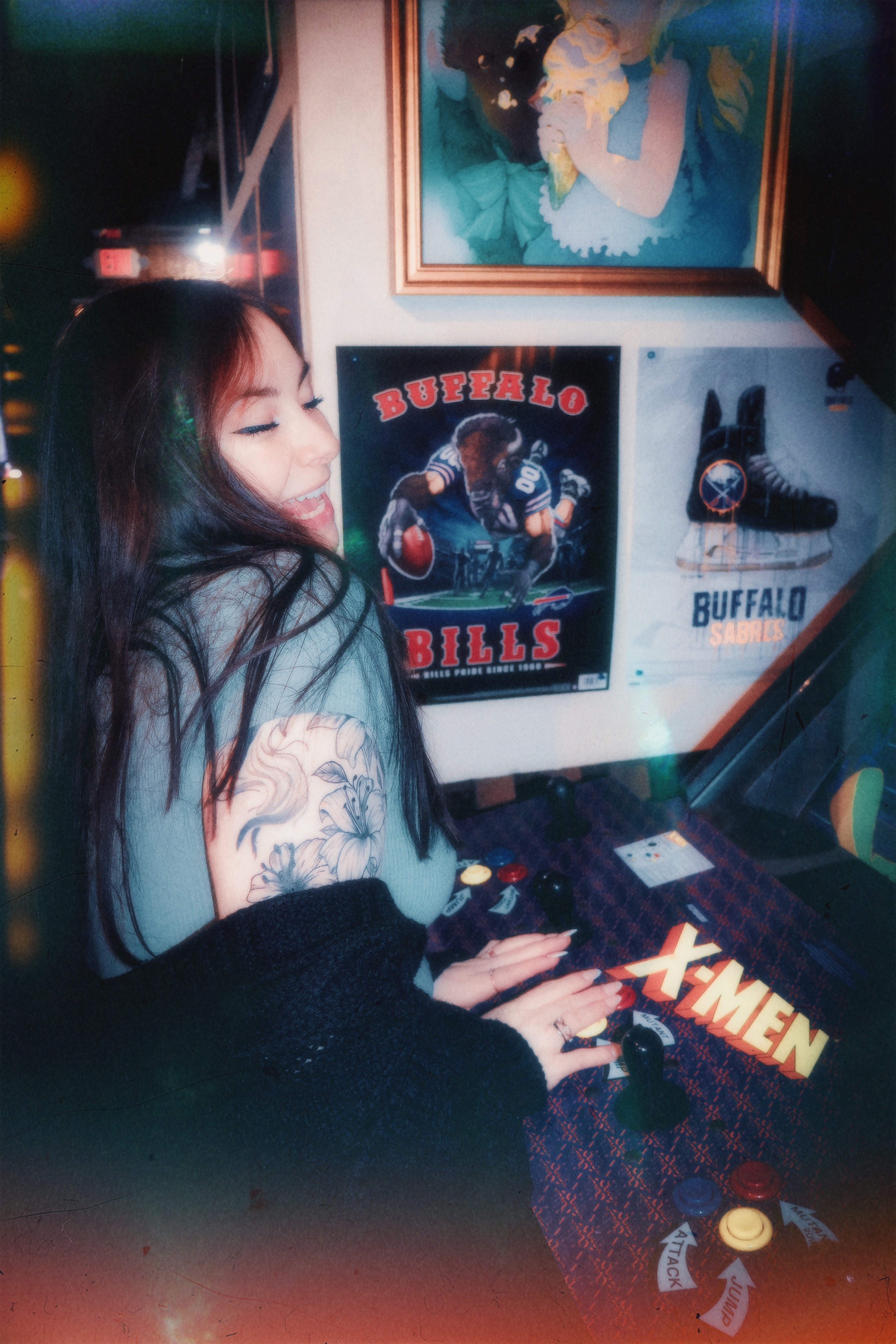 Person with long hair and a tattoo on arm, sitting at a gaming console, with posters and artwork on the wall behind.