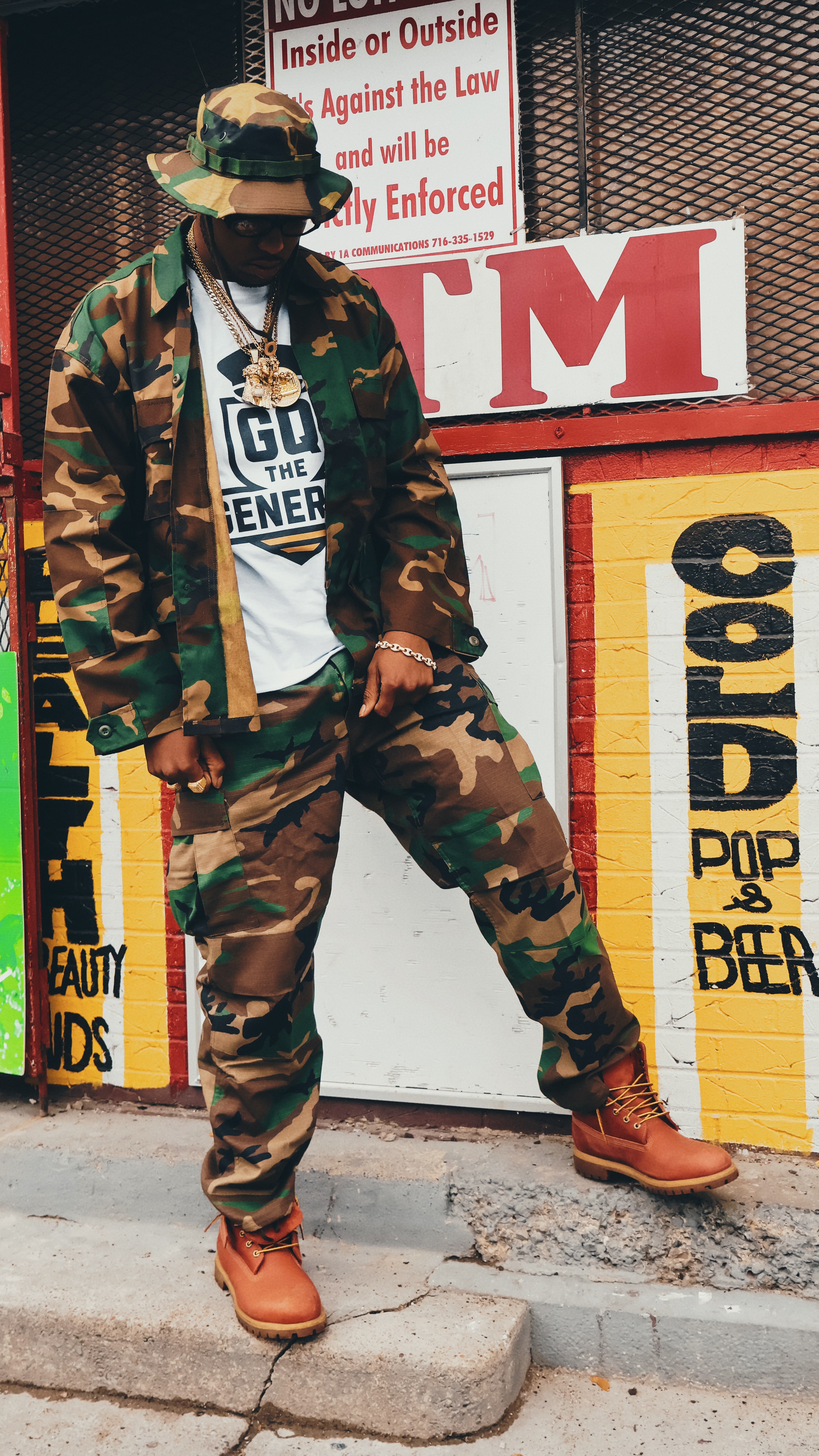 Person in camouflage outfit and boots standing outside near a colorful wall with signs and advertisements.