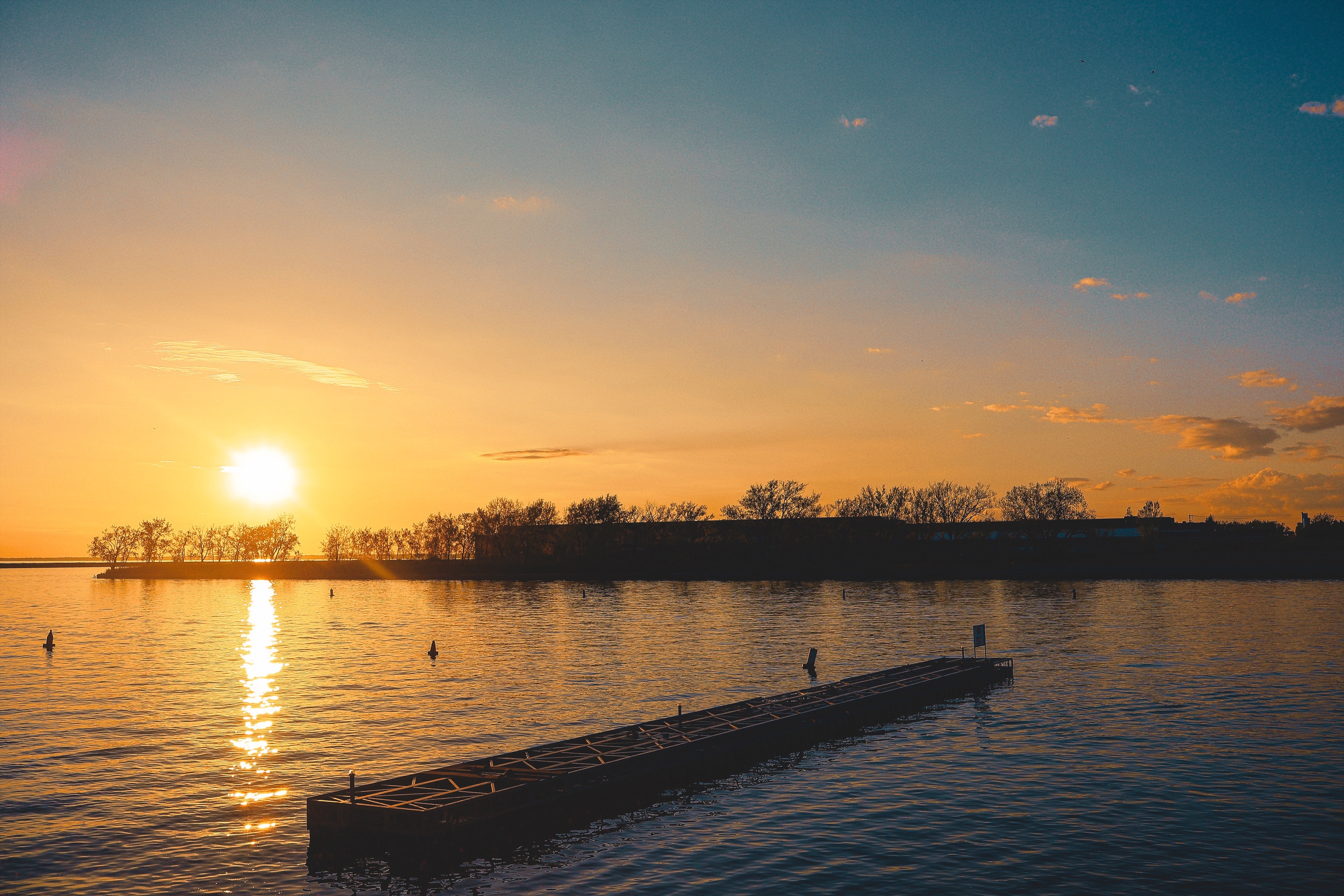 Sunset over a body of water with a dock and trees in the distance, sky with orange and blue hues.