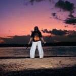 Person standing on beach facing water during sunset, holding a stick, wearing a black shirt with a fiery design and white pants.