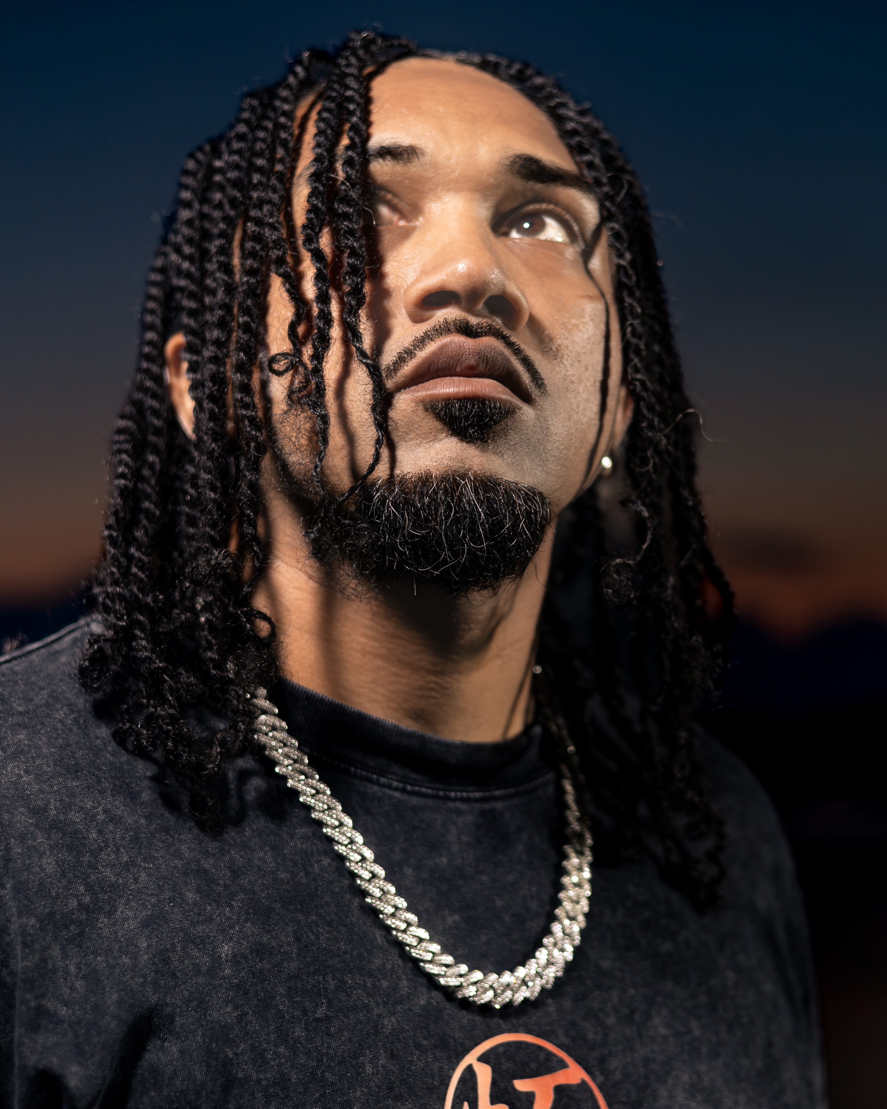 Man with braided hair and a chain necklace looks upward against a dark sky at sunset.