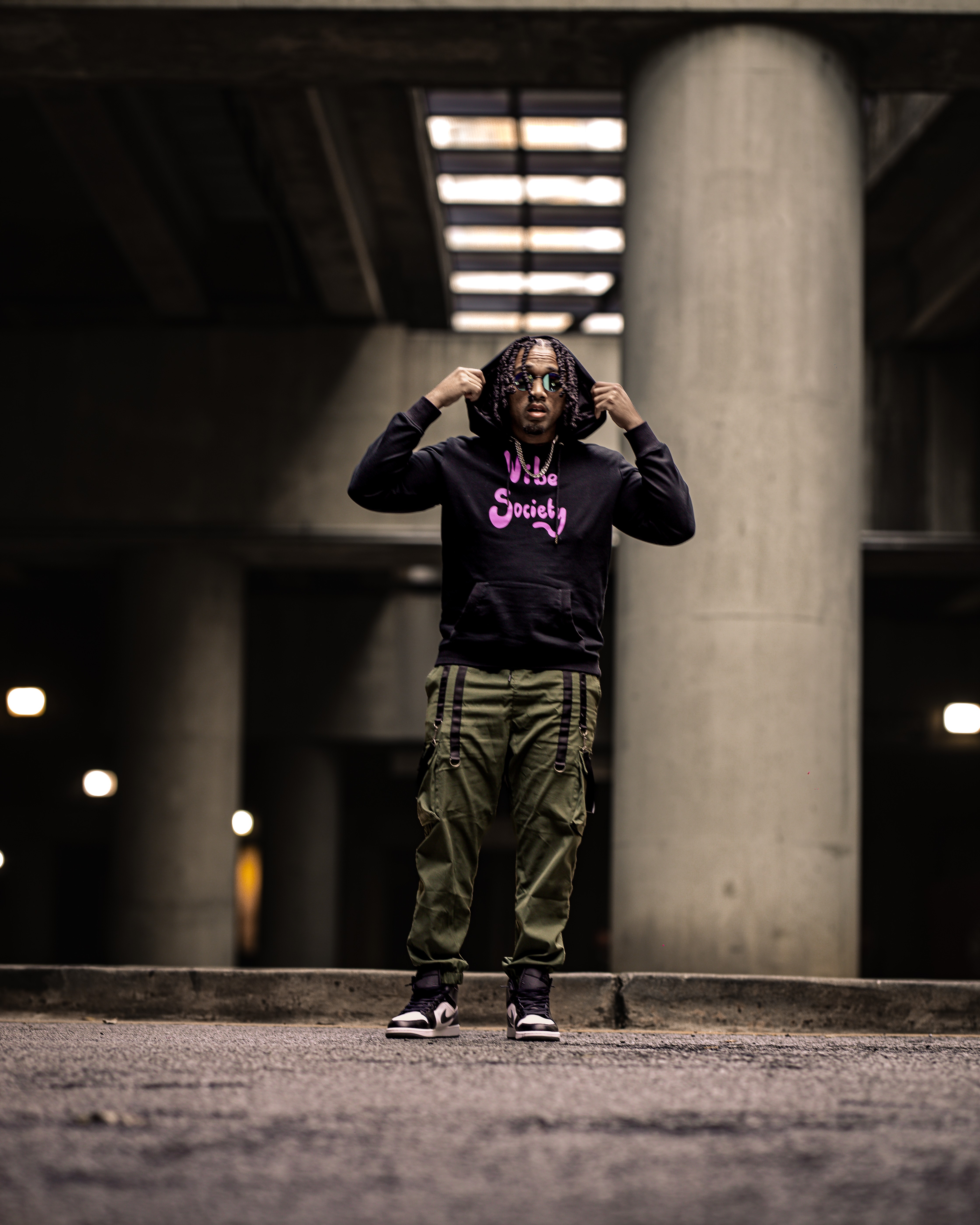 Person with curly hair wearing sunglasses, black hoodie with pink text, and camouflage pants standing under a concrete structure.