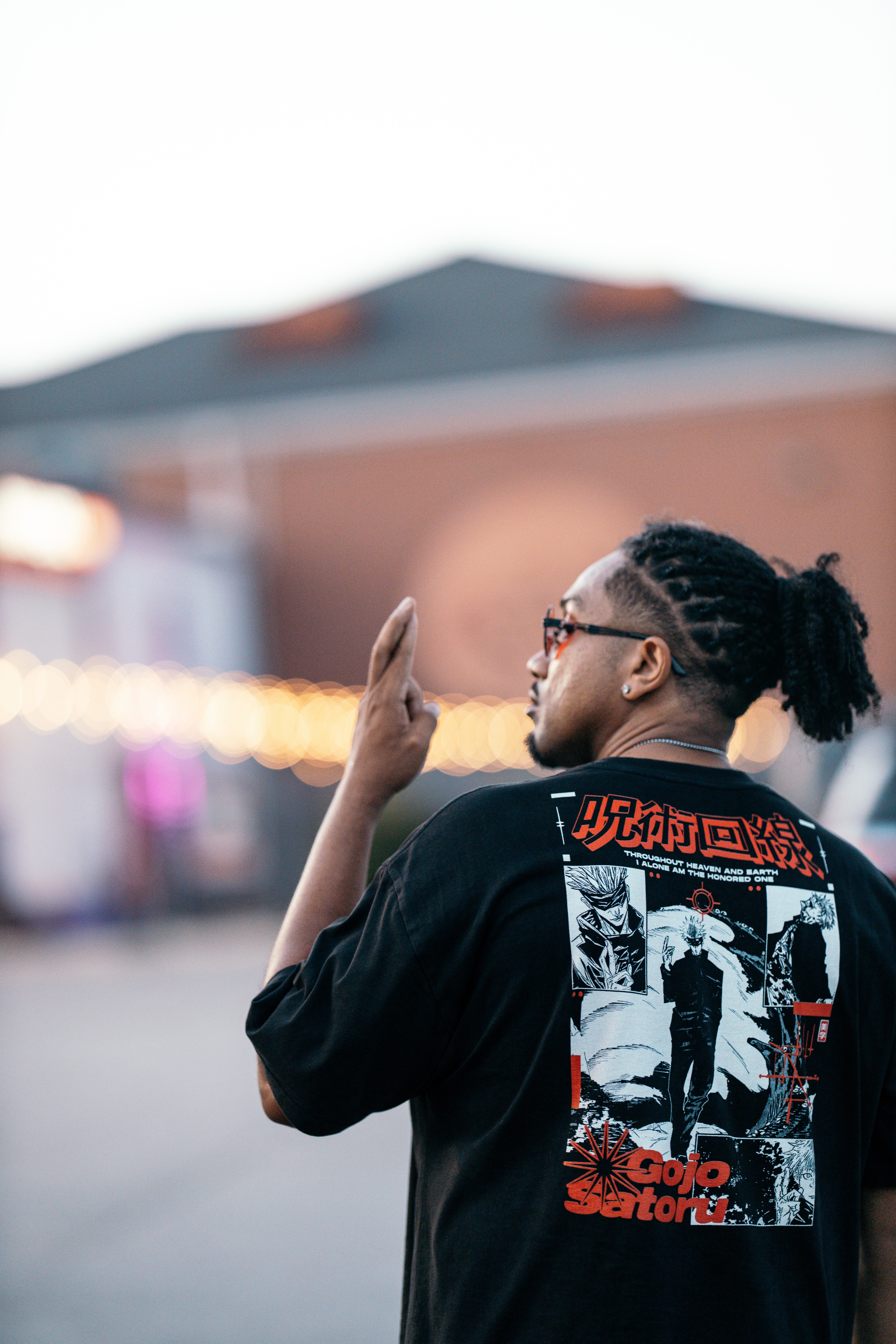 Person with glasses and braided hair, wearing a black graphic shirt, gesturing with hand outdoors.