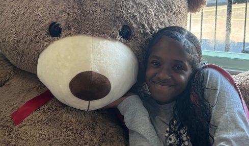 Person lying next to a large teddy bear with a white snout and black eyes, indoors near a window.