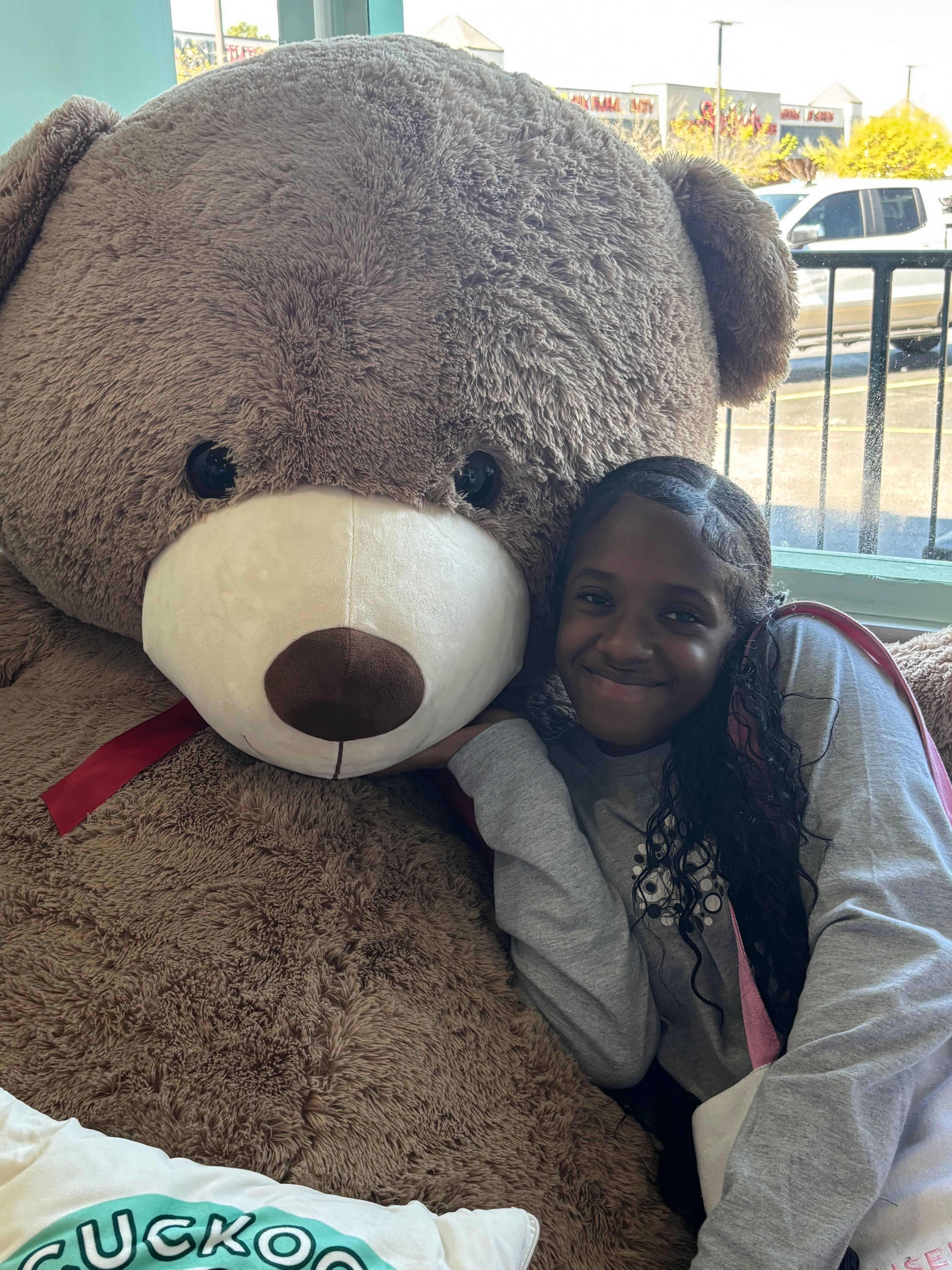 Person lying next to a large teddy bear with a white snout and black eyes, indoors near a window.