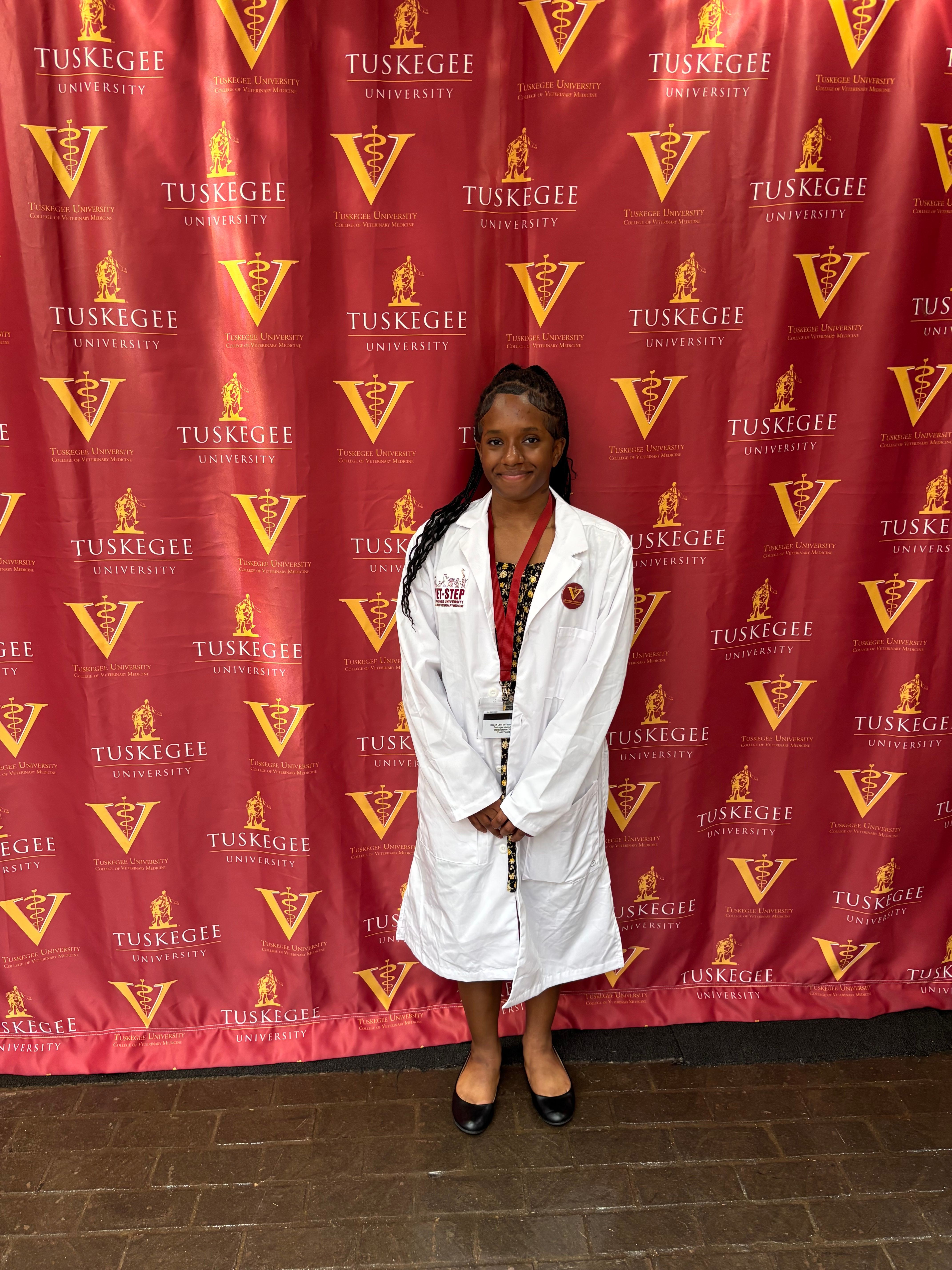 Woman in white coat and black shoes standing in front of red backdrop with yellow logos and text, smiling.